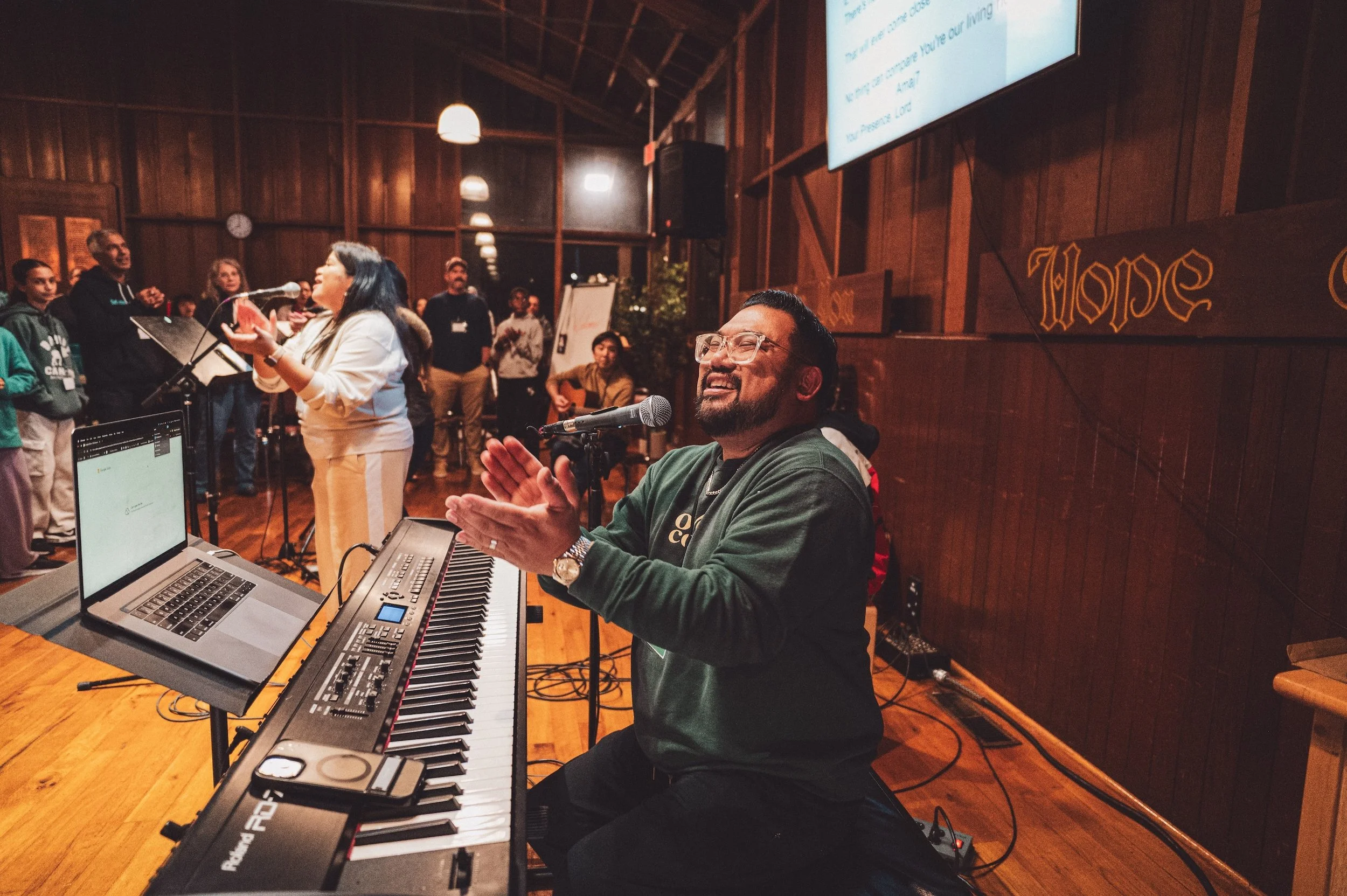 A man playing keyboard and singing into a microphone at an indoor event with a woman singing into a microphone and people gathered in the background.