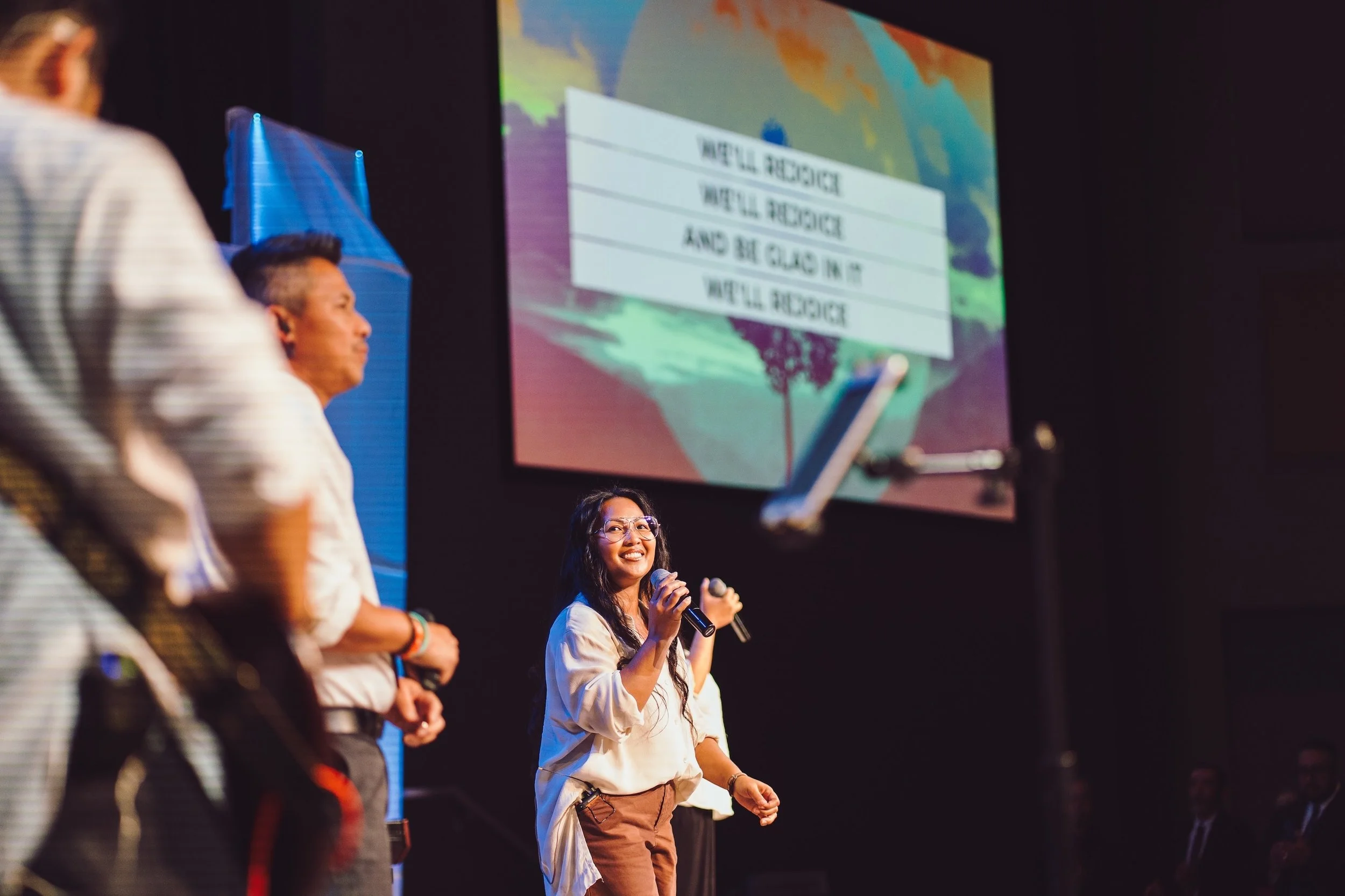 A woman with long dark hair and glasses holding a microphone, smiling while standing on a stage, with two men in the background and a screen displaying lyrics about making choices and being brave in the background.