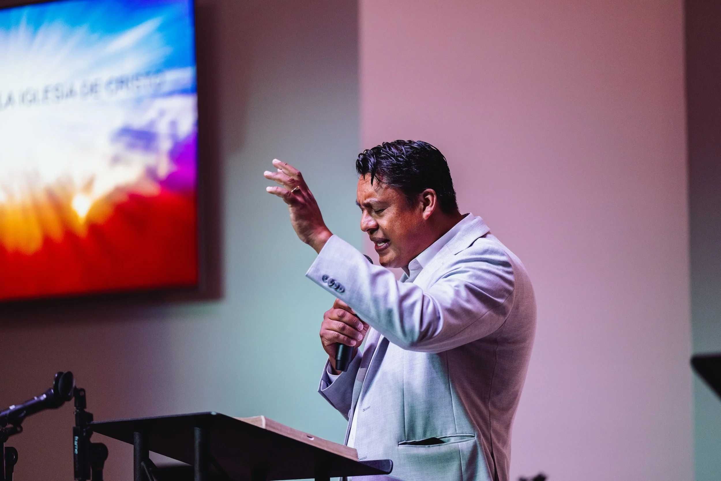 A man in a white suit speaking passionately at a podium with a microphone, gesturing with his hand, in front of a large screen displaying colorful graphics.