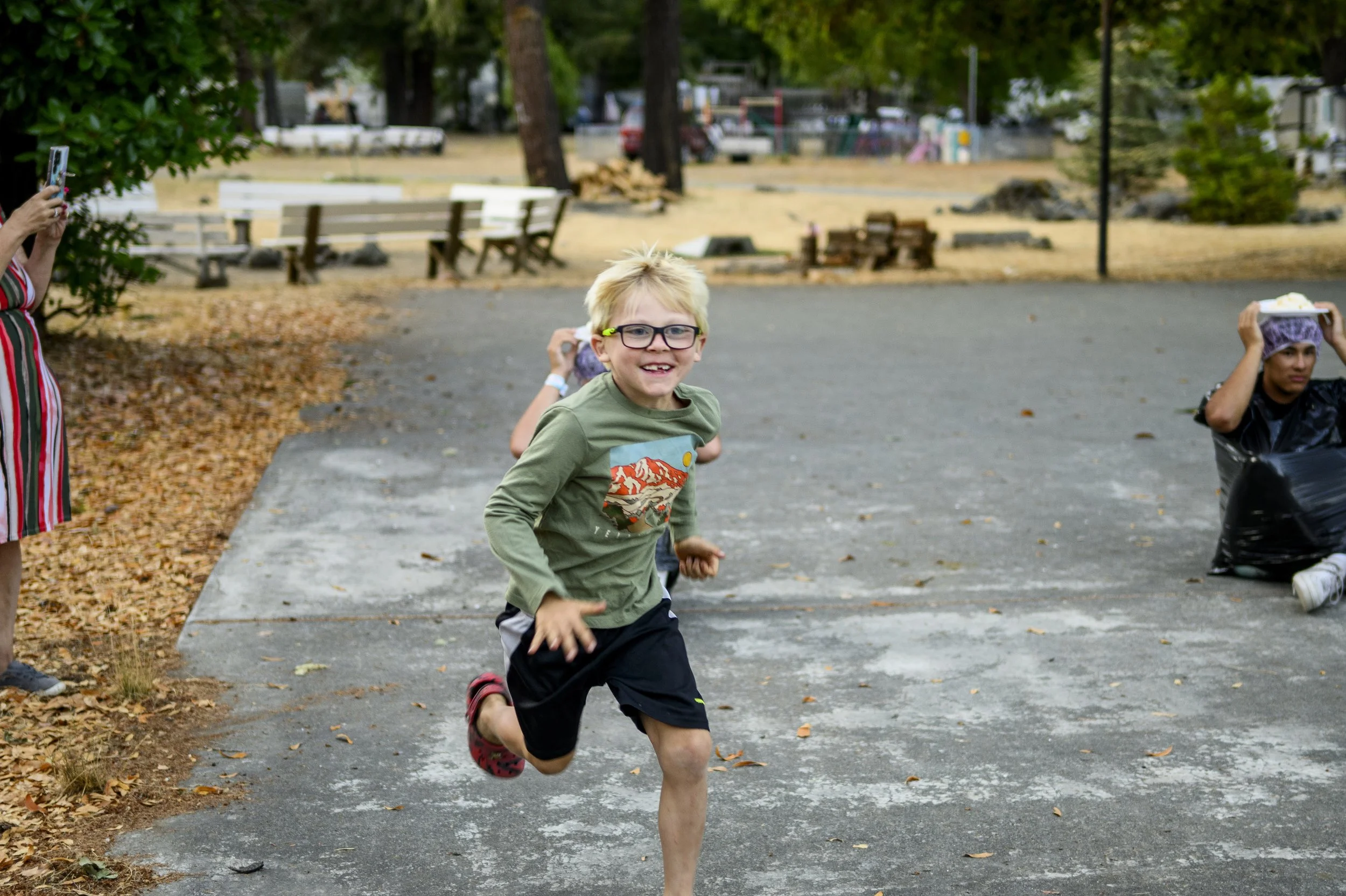 A young boy running and smiling outdoors in a park, with people taking photos nearby and park benches in the background.