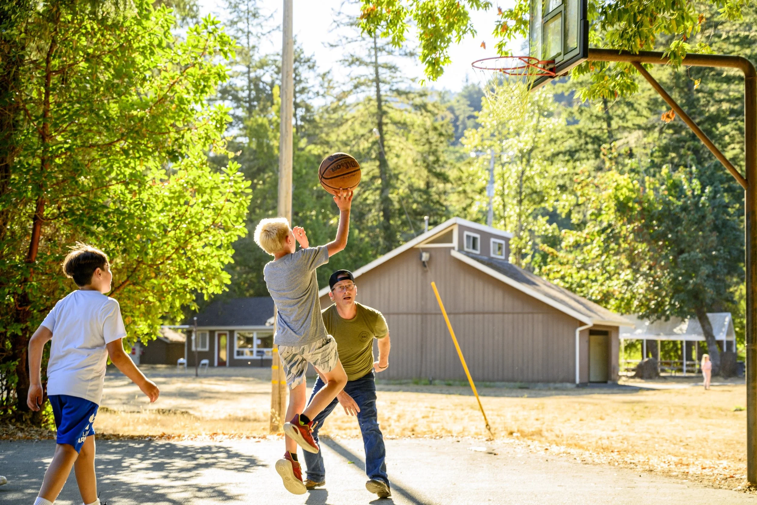 Three children and an adult playing basketball outdoors on a sunny day. One child is jumping to make a shot while the adult is trying to block, and another child watches. There are trees and a house in the background.
