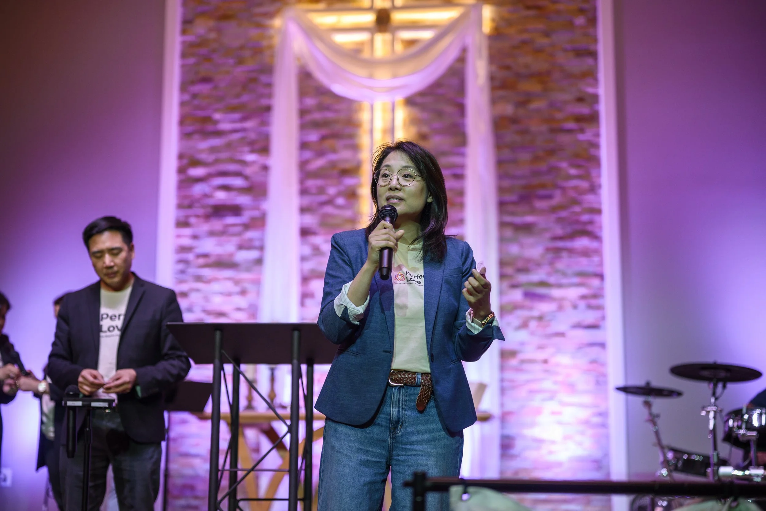 A woman wearing glasses and a blue blazer speaks into a microphone on stage, with a backdrop of a brick wall and draped fabric, accompanied by a man in a suit and others in the background, at an indoor event.