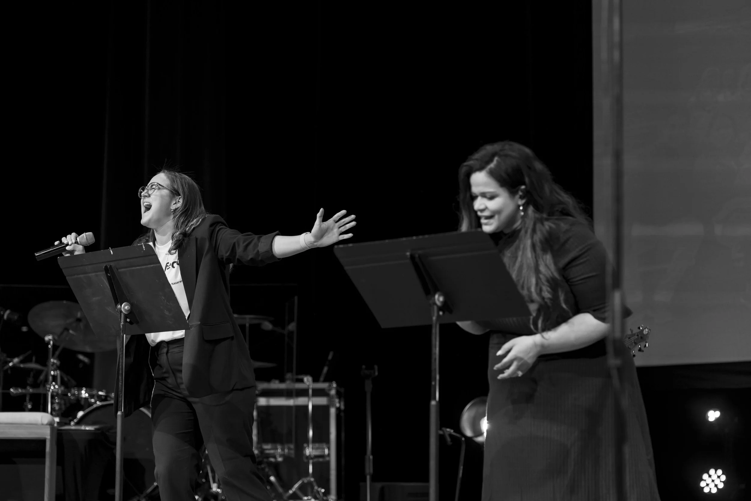 Two women singing passionately on stage, one holding a microphone and expressive with her arms outstretched, the other with a music stand in front of her, both appearing joyful during a performance.