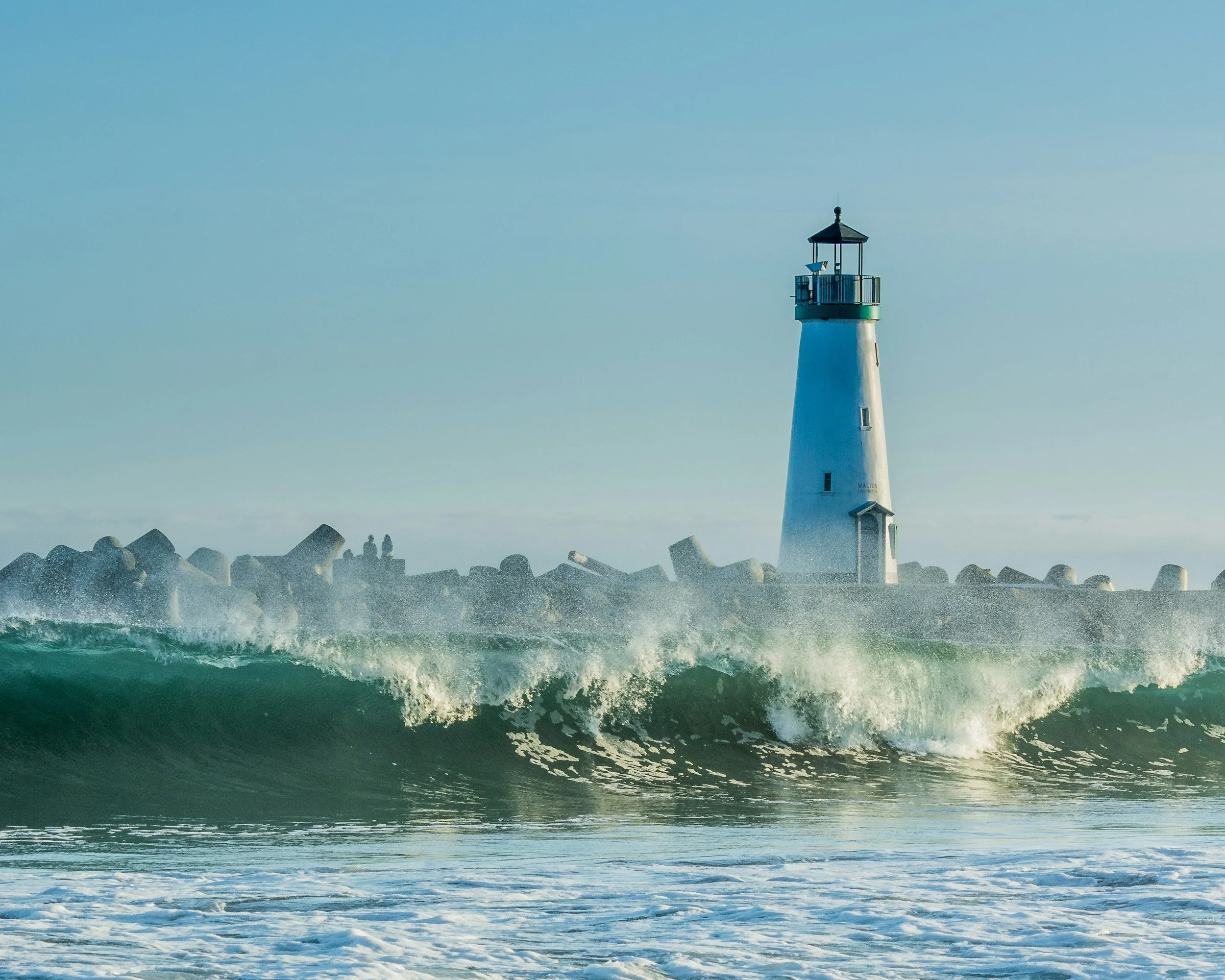 Lighthouse on a rocky breakwater with rough ocean waves in the foreground and a clear sky.
