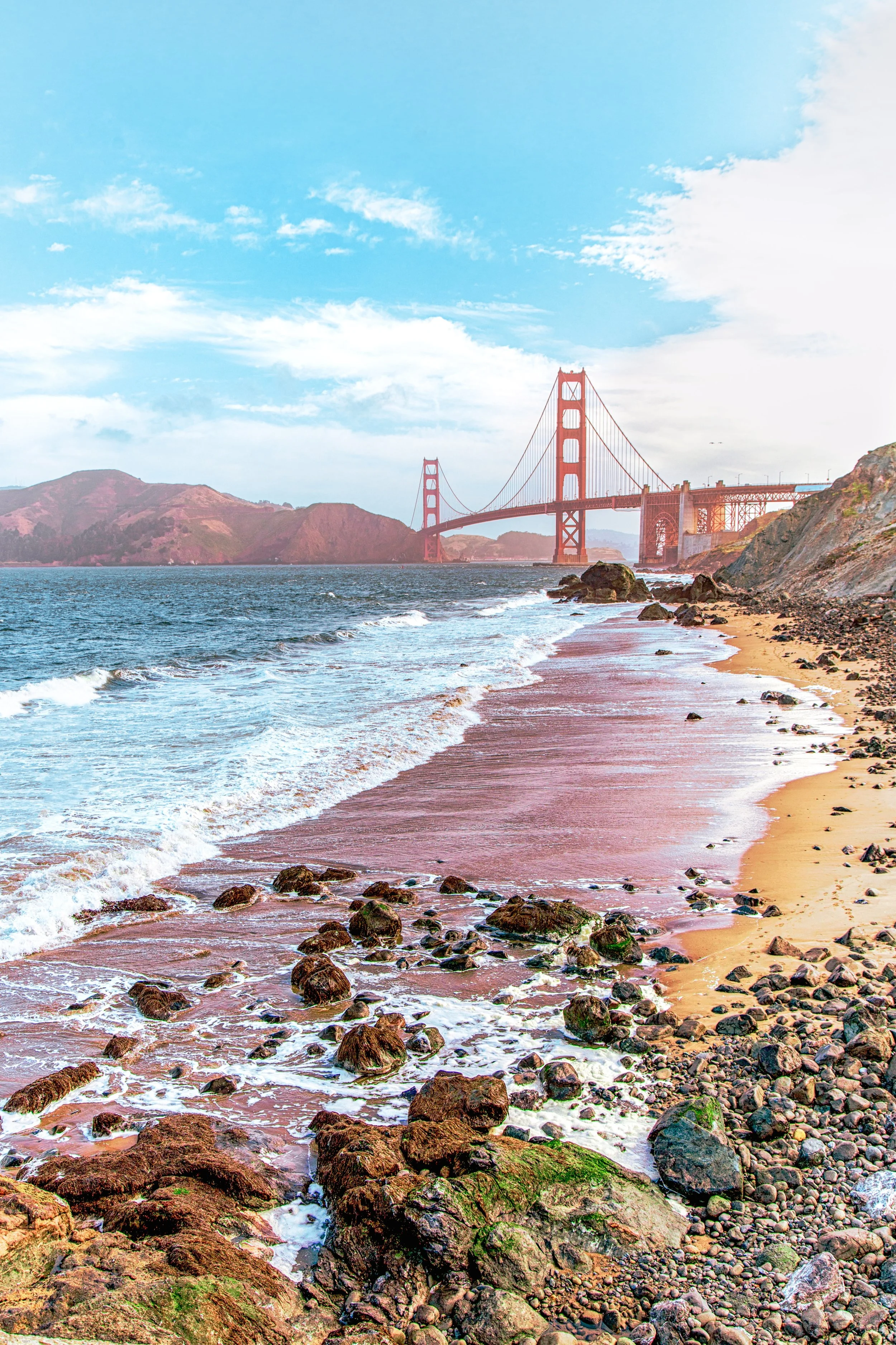 View of the Golden Gate Bridge over the San Francisco Bay with rocks and sandy beach in the foreground, under a partly cloudy sky.