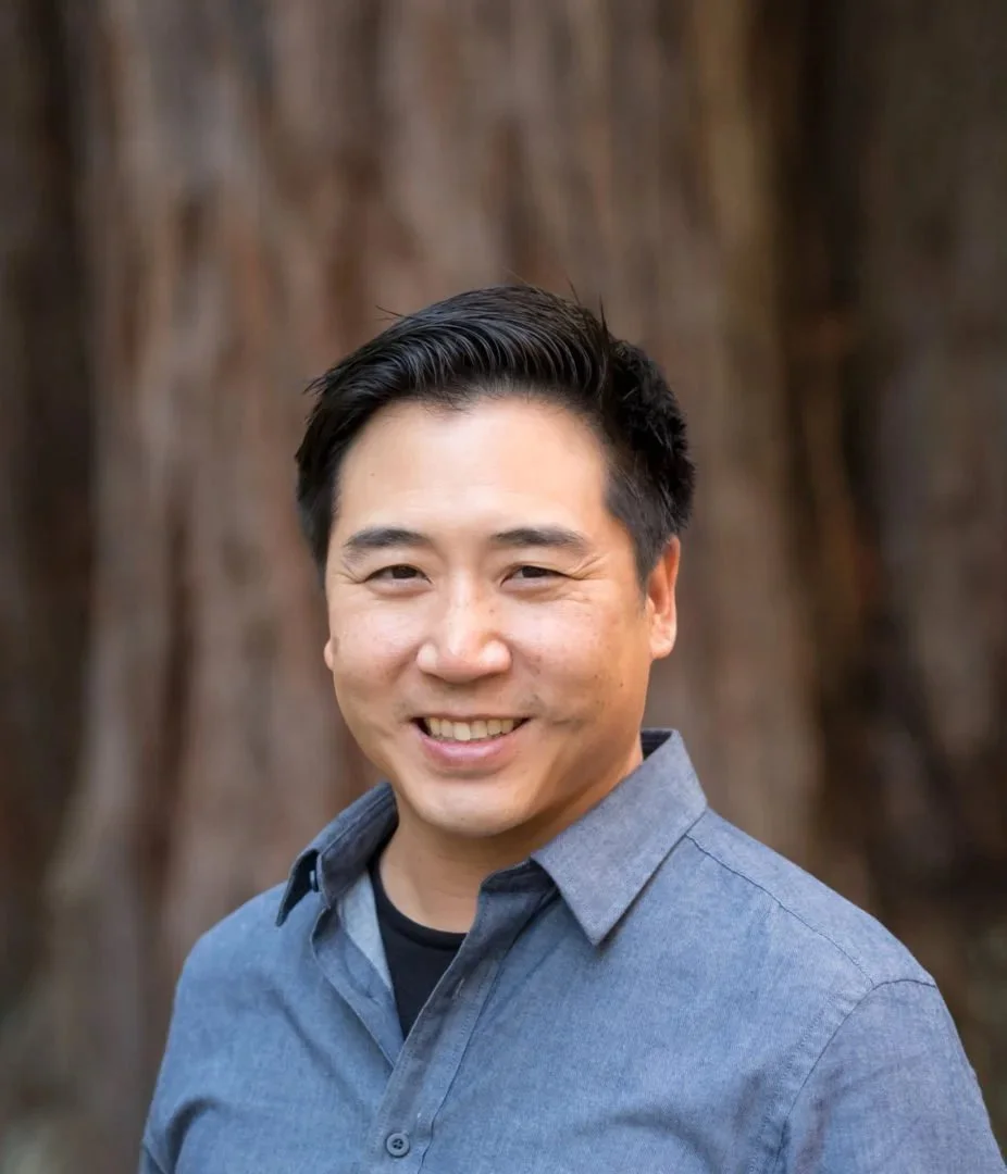 Smiling man with short black hair, wearing a blue button-up shirt, standing outdoors with large tree trunk in the background.
