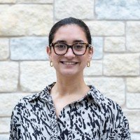 A woman with glasses and dark hair tied back, smiling, wearing a black and white patterned blouse, standing outdoors against a stone wall background.