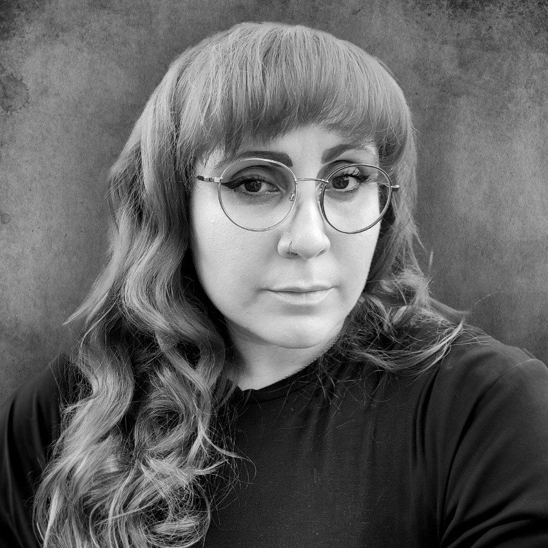 A woman with wavy hair wearing glasses and a dark top, looking at the camera against a plain background.