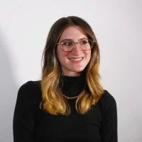 A woman with shoulder-length brown hair, wearing glasses and a black top, smiling against a white background.