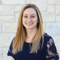 Portrait of a young woman with long blonde hair, smiling, wearing a navy blue top with lace sleeves, standing against a light stone wall.