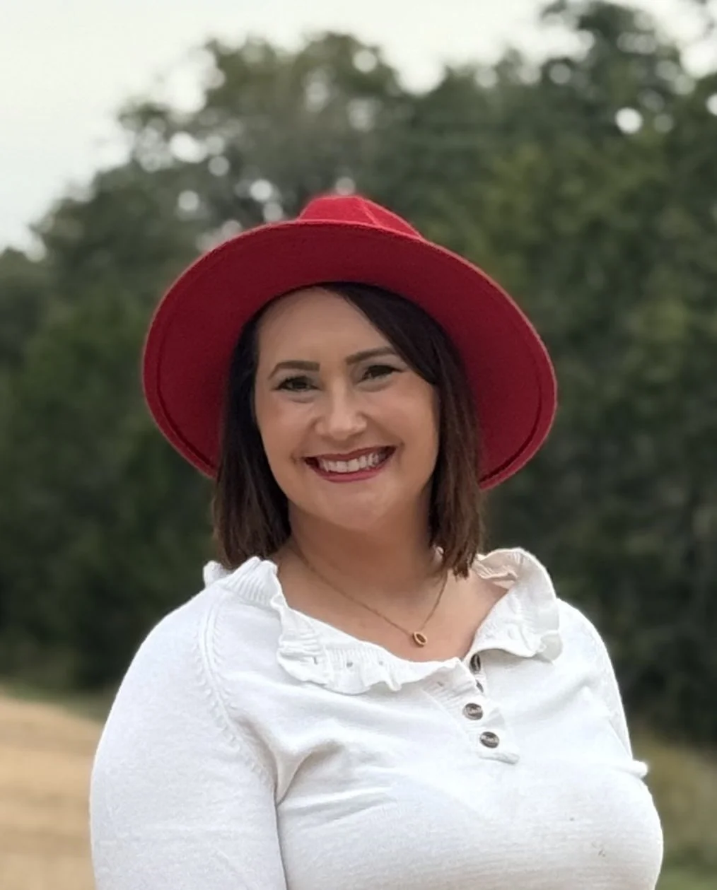 A woman smiling, wearing a red hat and a white shirt, outdoors with trees in the background.