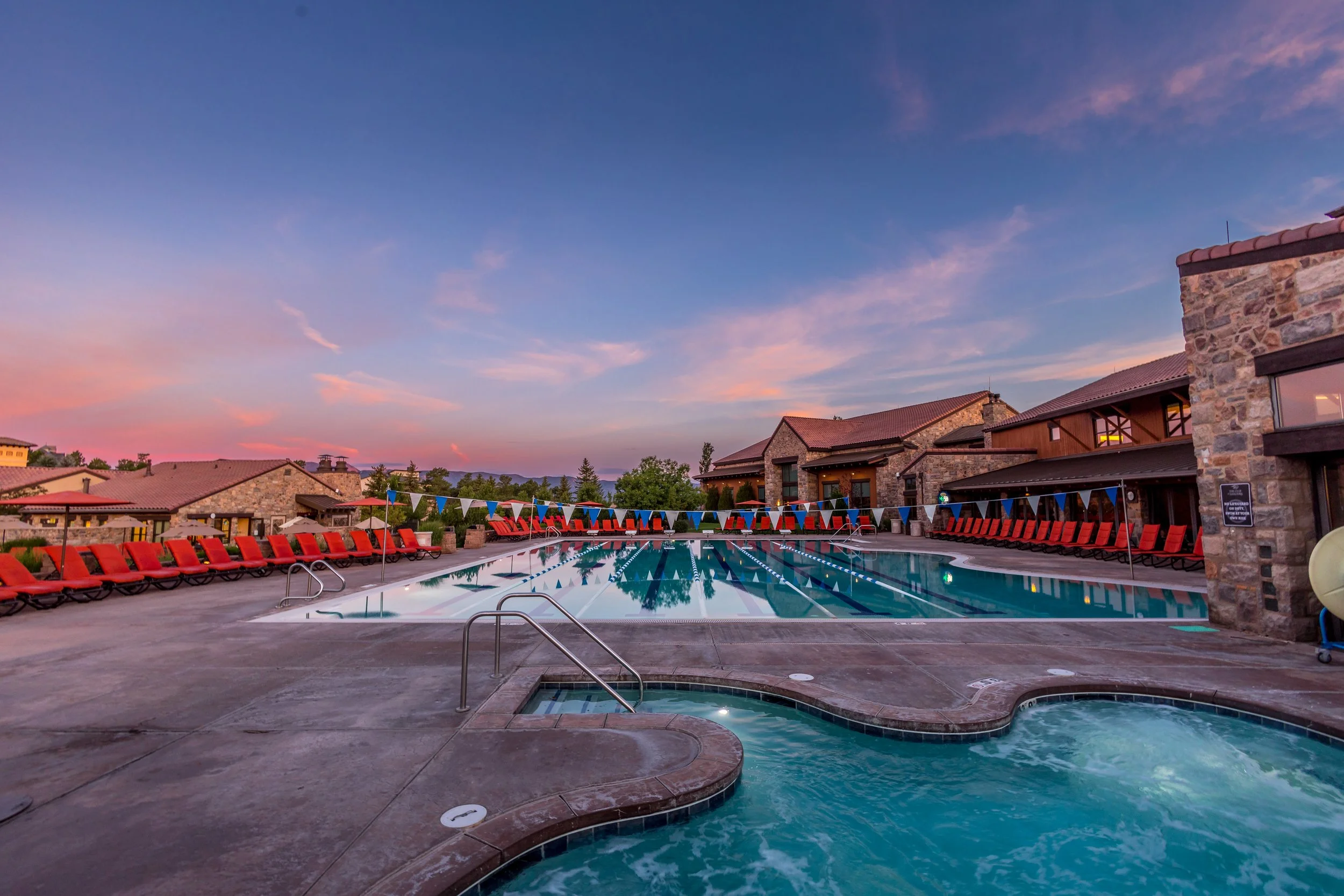An outdoor swimming pool and hot tub area at sunset with empty red lounge chairs lining the pool and a stone building in the background.