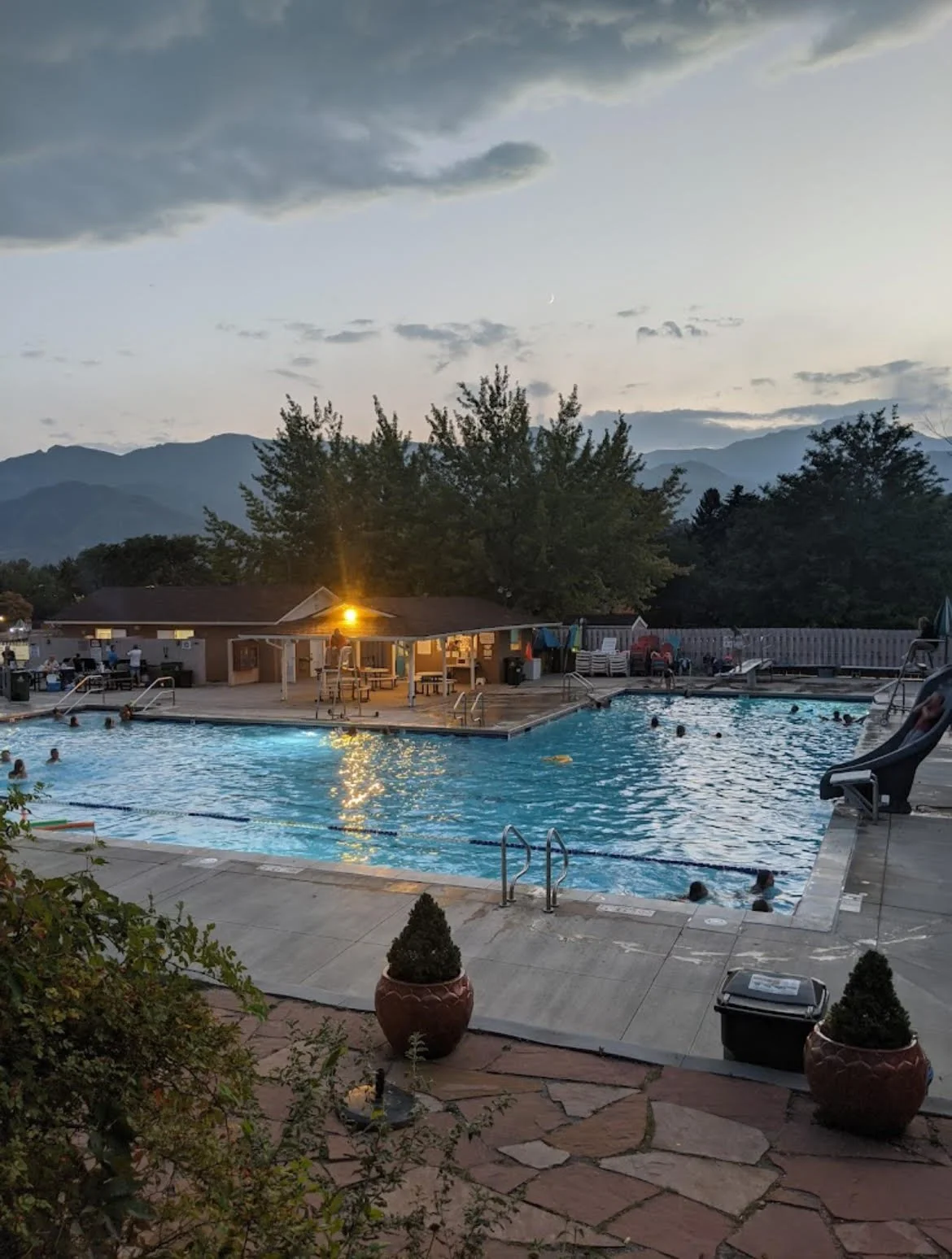 An outdoor swimming pool scene at dusk with people swimming, a slide, trees, mountains in the background, and a fenced area with lounge chairs and tables, illuminated by an outdoor light.