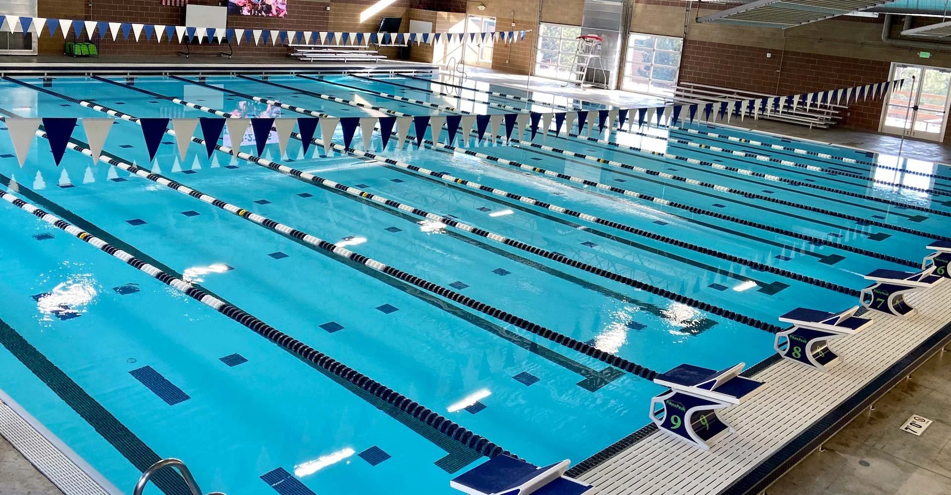 Indoor swimming pool with multiple lanes separated by black and white lane dividers, blue and white triangular flags hanging overhead, starting blocks labeled with lane numbers, and large windows letting in natural light.
