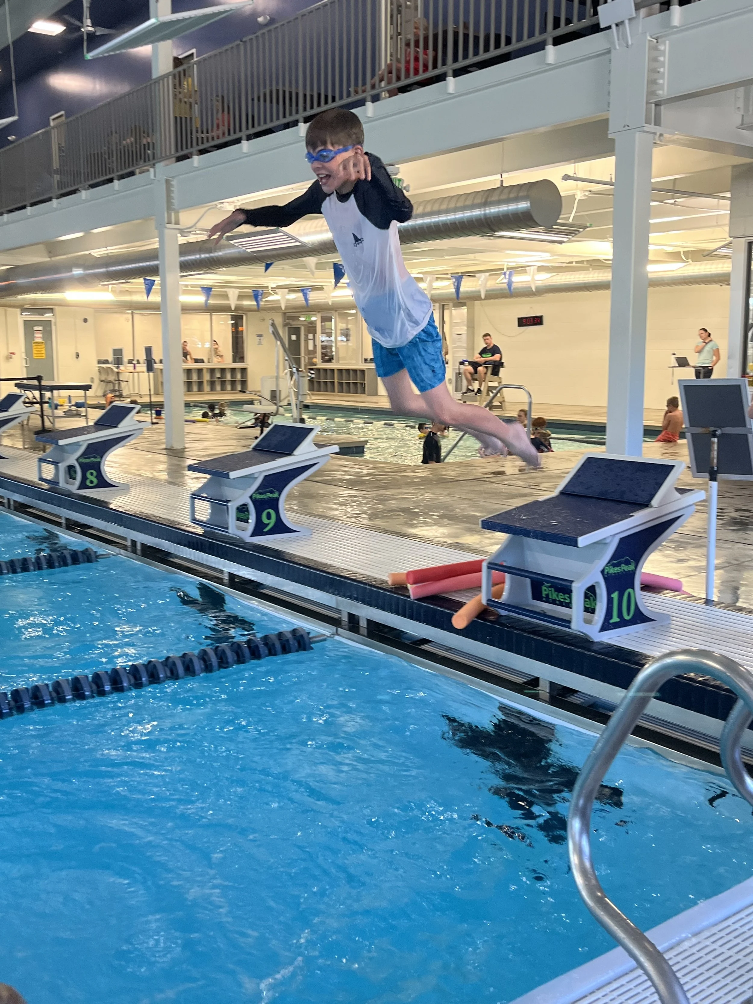 A young boy wearing swim goggles, a white and black long-sleeved rash guard, and blue swim shorts is jumping off the starting block into an indoor swimming pool, with several people in the background.