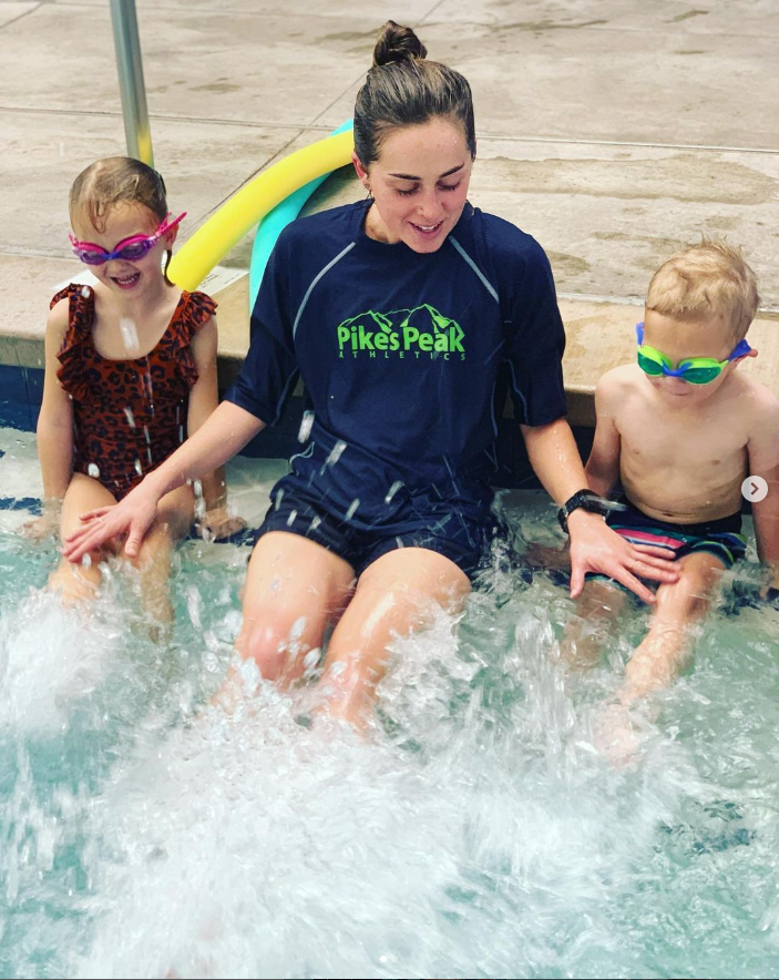 A woman and two children sitting at the edge of a swimming pool, splashing water with their feet. The woman is wearing a navy t-shirt with 'Pikes Peak Athletics' printed on it. The girl on the left is wearing a leopard print swimsuit and pink goggles, while the boy on the right is shirtless, wearing striped swim trunks and green goggles.