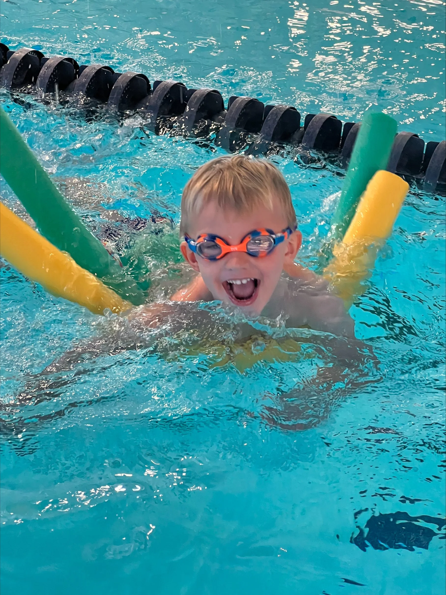 A young boy with swimming goggles smiling happily in a swimming pool, surrounded by colorful pool noodles.