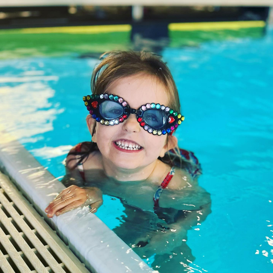 A young girl smiling in a swimming pool, wearing colorful, rhinestone-encrusted sunglasses shaped like butterflies.