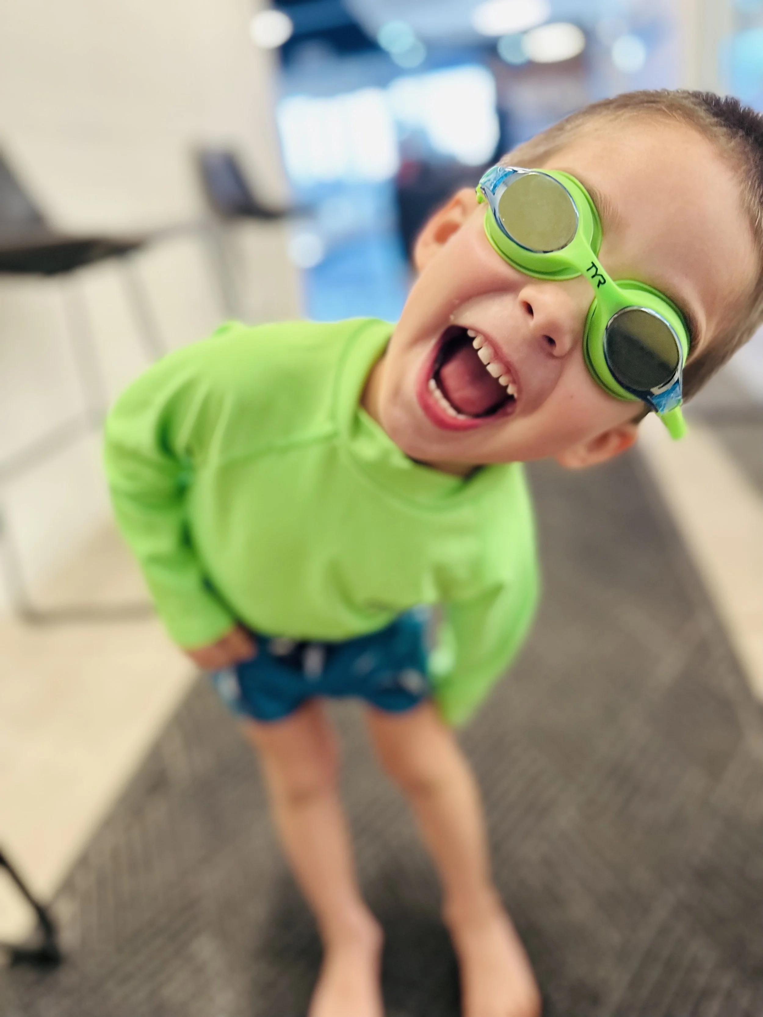 A young boy wearing bright green swim goggles and a neon green jacket, smiling and leaning towards the camera in an indoor setting.
