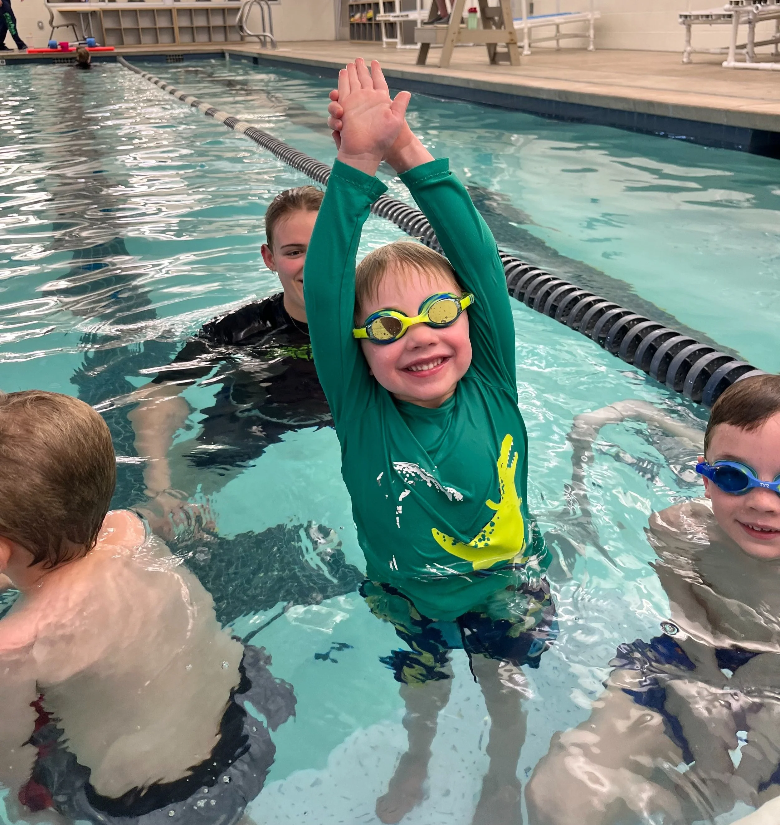 Children swimming in a pool, with one young boy wearing green goggles and a green shirt smiling with arms raised, surrounded by other kids swimming.