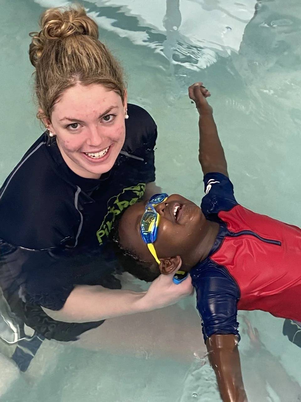 A woman assists a child in the water, helping them float on their back during swimming practice, both smiling and wearing goggles.