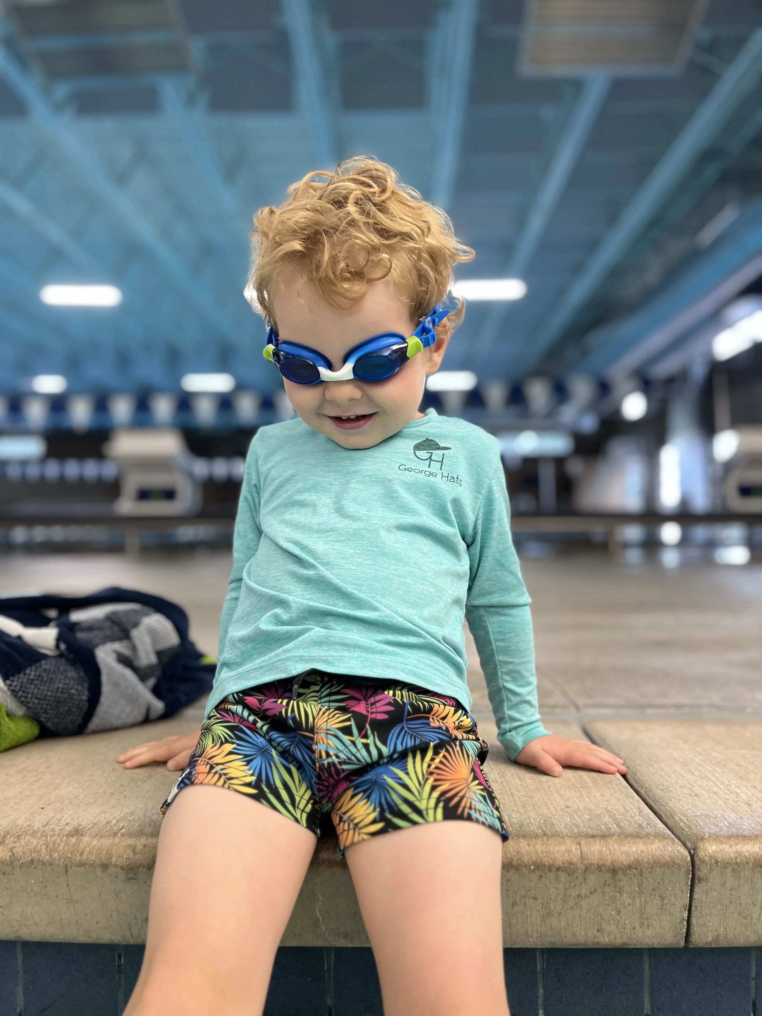Young boy with curly red hair wearing blue swim goggles, a long-sleeve teal shirt with a logo, and colorful tropical swim trunks, sitting on a bench at an indoor swimming pool.