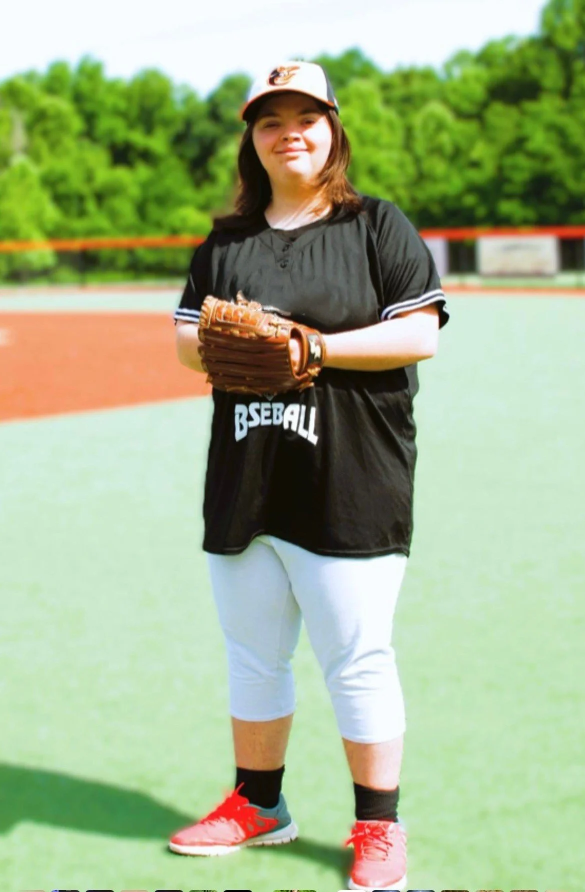 Young woman in baseball uniform standing on a field, holding a baseball glove, with trees and a baseball diamond in the background.