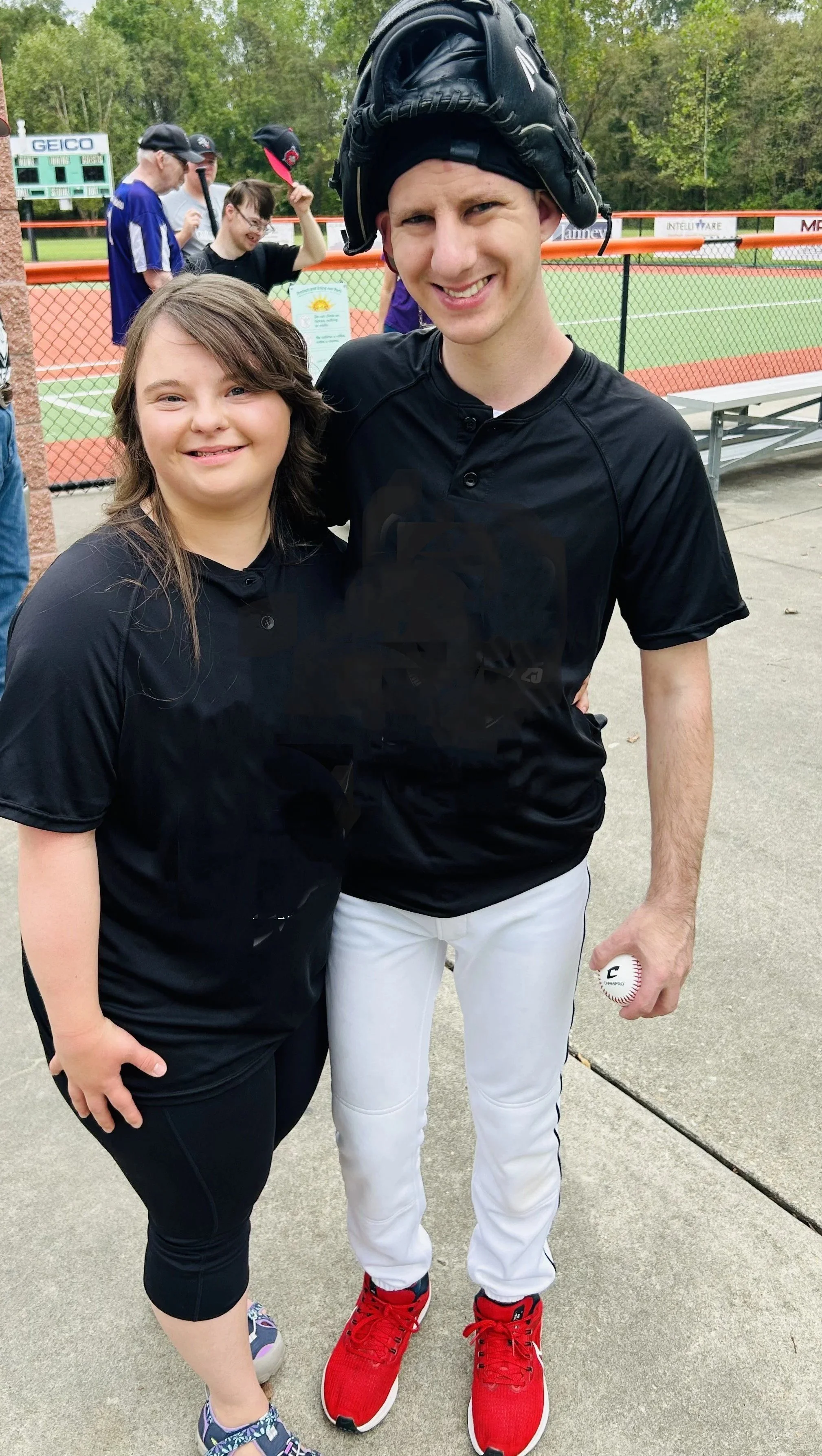 A young girl and a male baseball player pose together at a baseball field, smiling at the camera. The player is wearing a black shirt, white pants, red shoes, and a baseball glove on his head, holding a baseball in his right hand. The girl is wearing