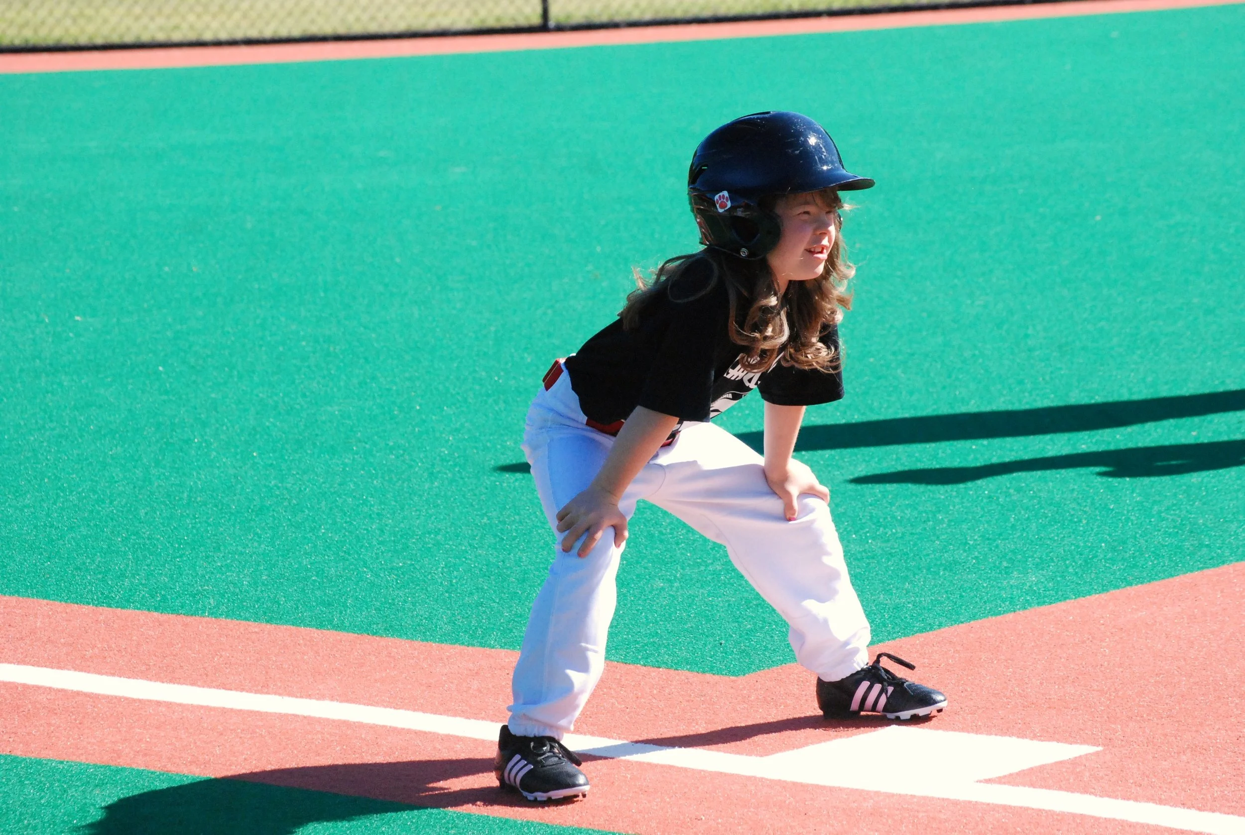 A young girl wearing a black helmet, black t-shirt, white pants, and black sneakers is in a ready position on a baseball or softball field with green and pinkish turf.