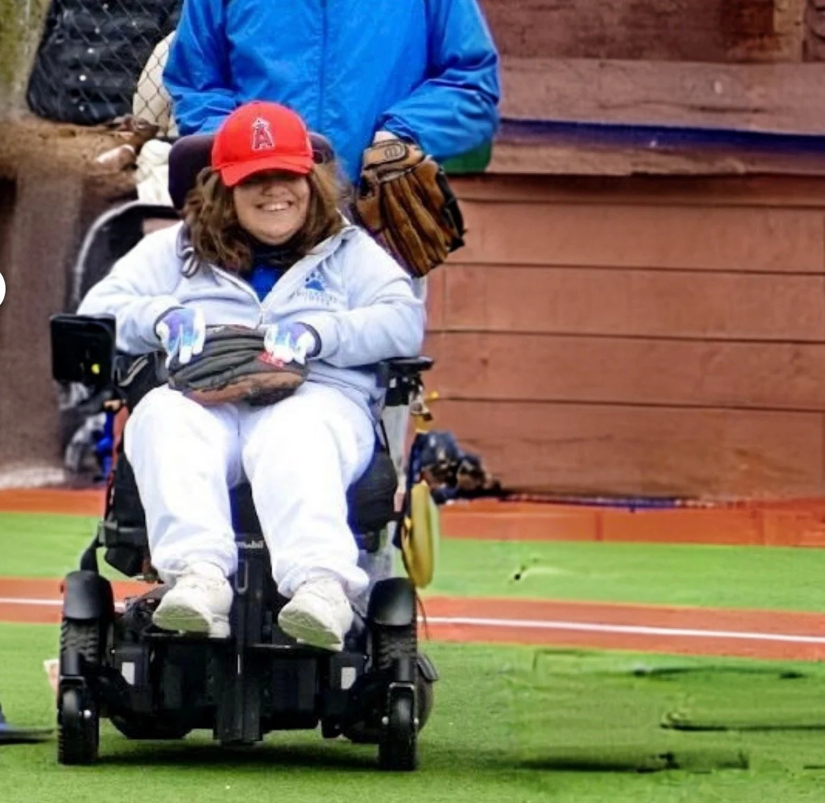 Person in a motorized wheelchair smiling at a baseball game, wearing a red cap, white jacket, and white pants, with baseball gloves on hands. Someone standing behind with a blue jacket and glove, on a baseball field.
