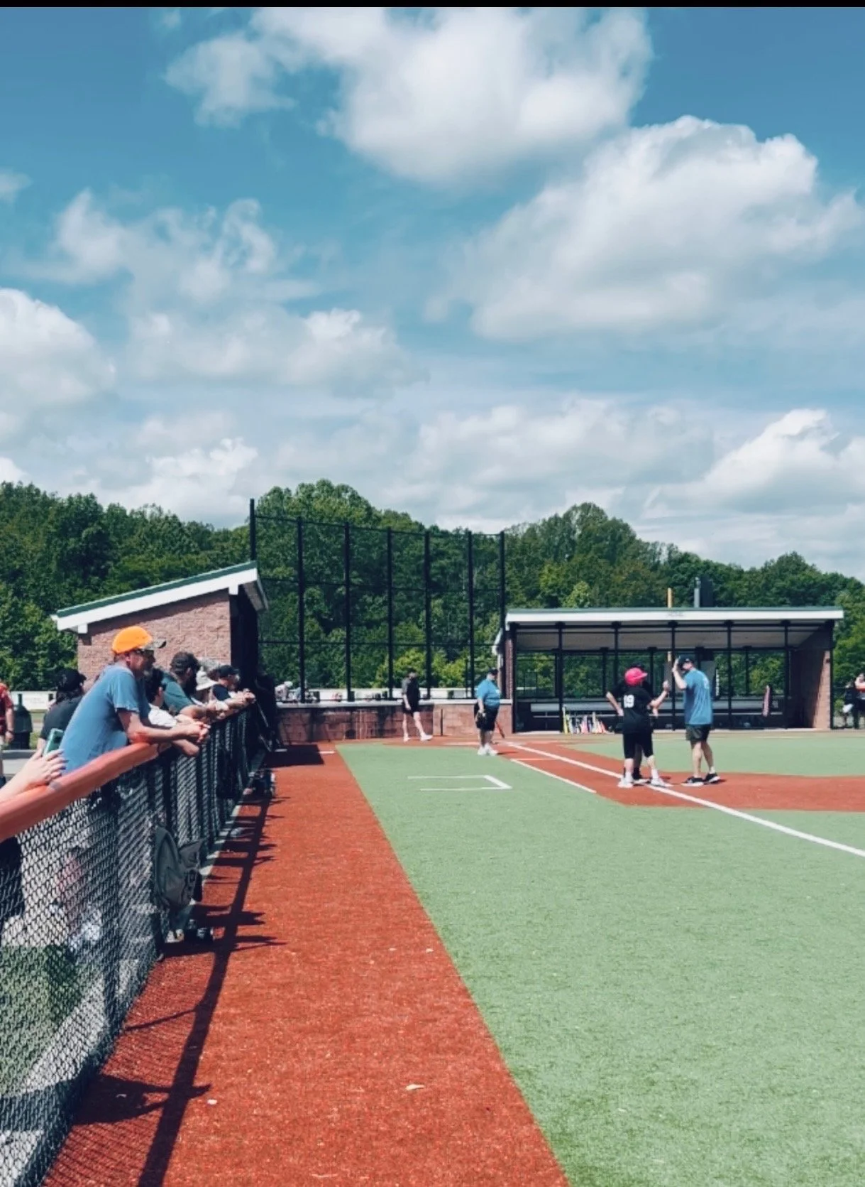 People watching a youth baseball or softball game at a field with a chain-link fence, trees, and cloudy sky in the background.
