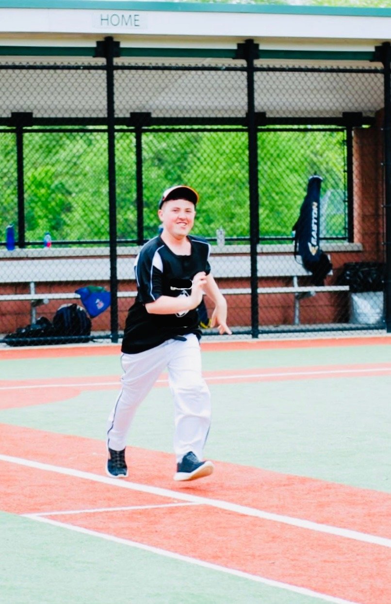 A boy running on a tennis court with a smile, dressed in a black sports shirt, white pants, and black shoes. There are tennis bags and bottles in the background.