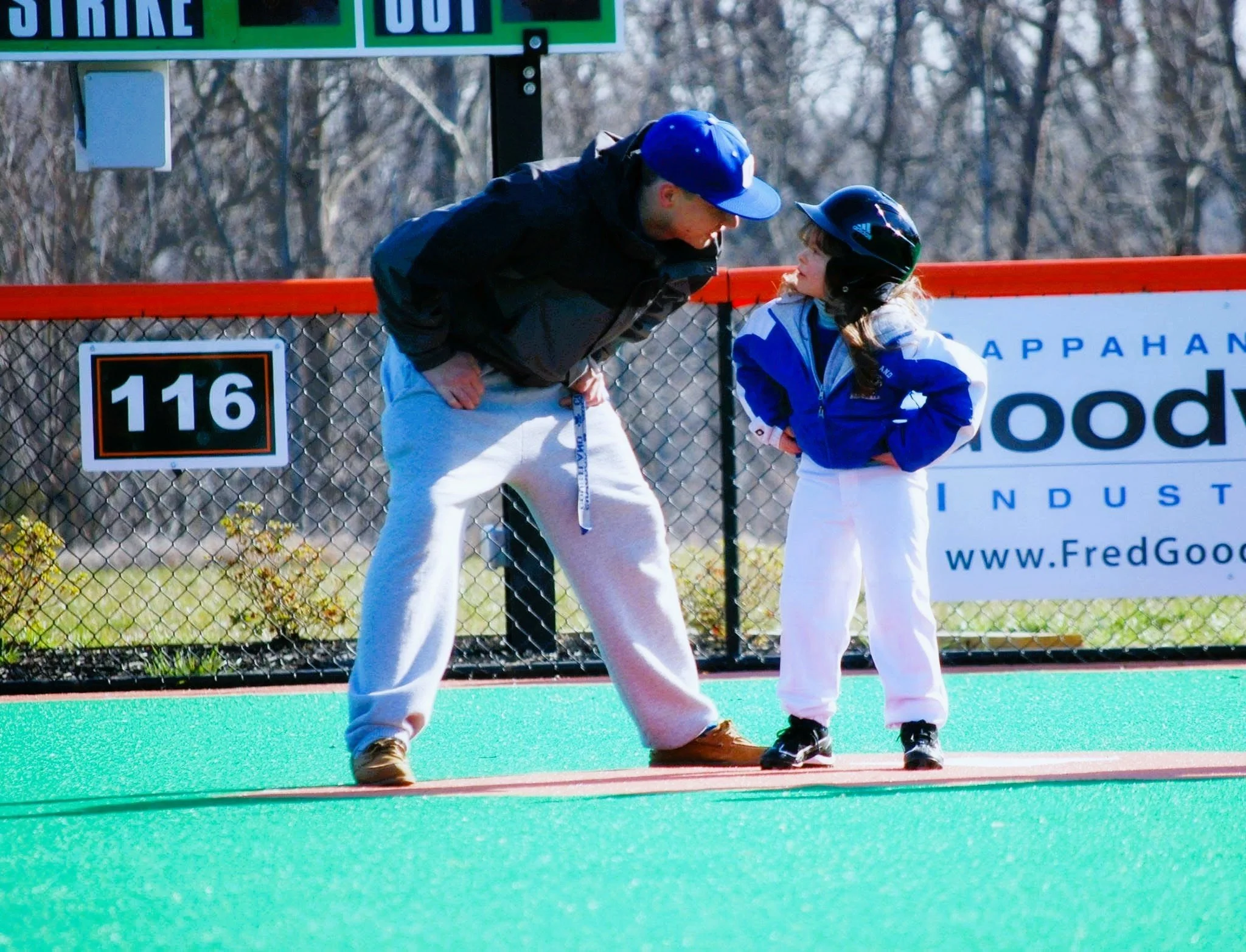 A man and a girl, both wearing winter clothing and helmets, are standing on a green sports field, engaged in a conversation or coaching moment.