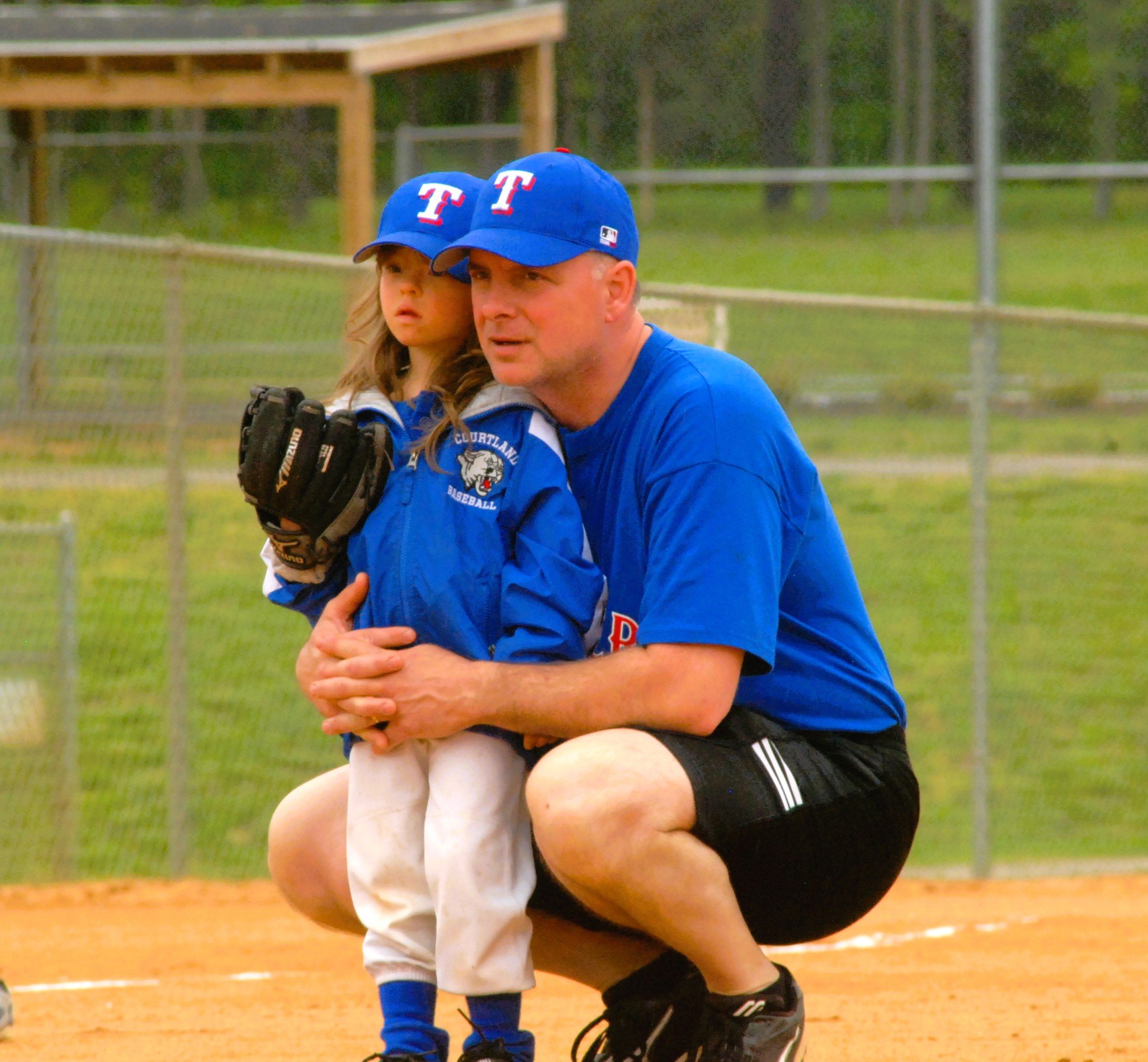 A father kneels on a baseball field, comforting his young daughter who is wearing a blue jacket, white pants, and a BlueT baseball cap, with a baseball glove on her left hand, during a youth baseball game.