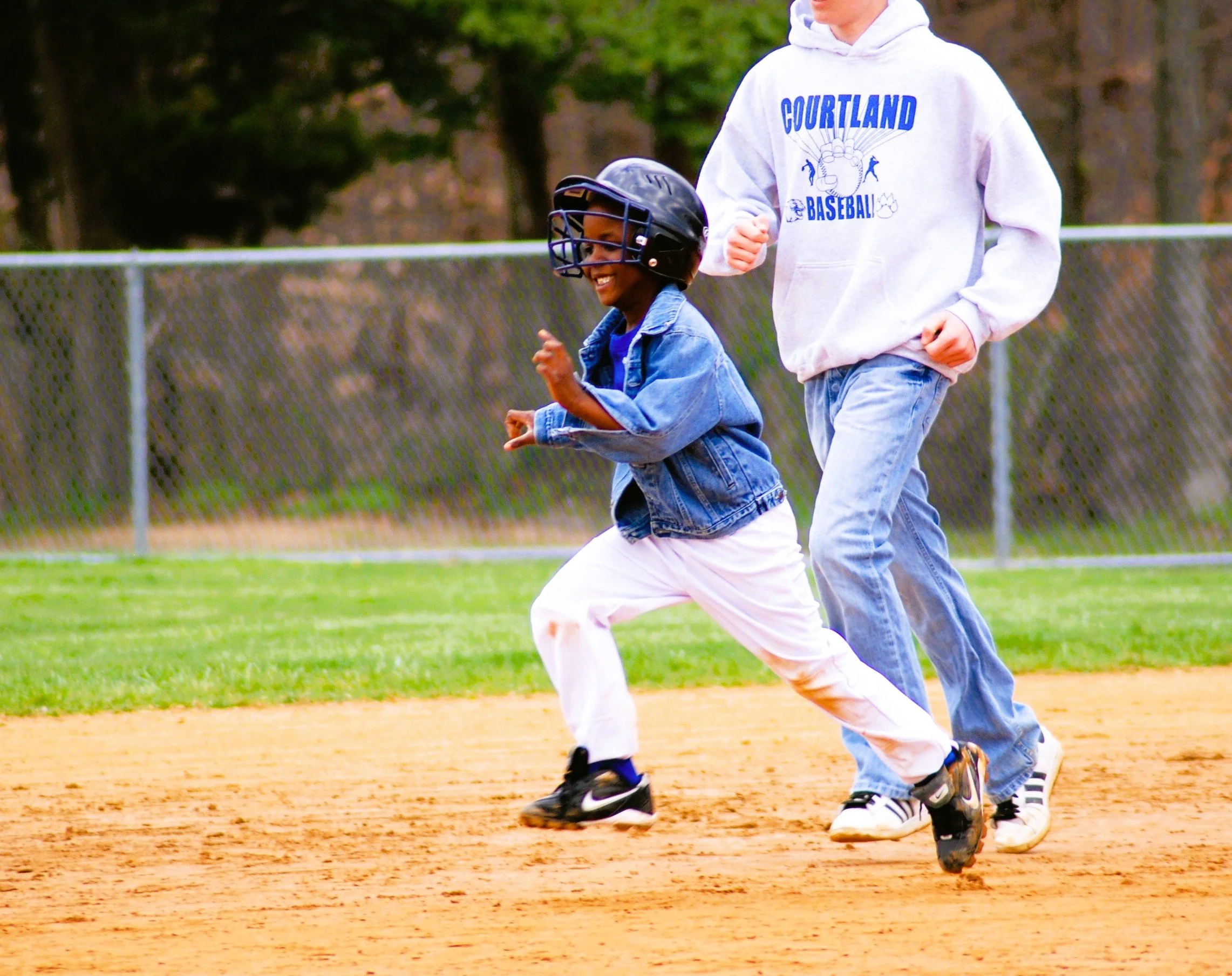 Two children running on a baseball field. The child in the foreground is wearing a black helmet, denim jacket, white pants, and black sneakers, smiling. The other child, partially visible, is wearing a white hoodie with blue lettering and light jeans
