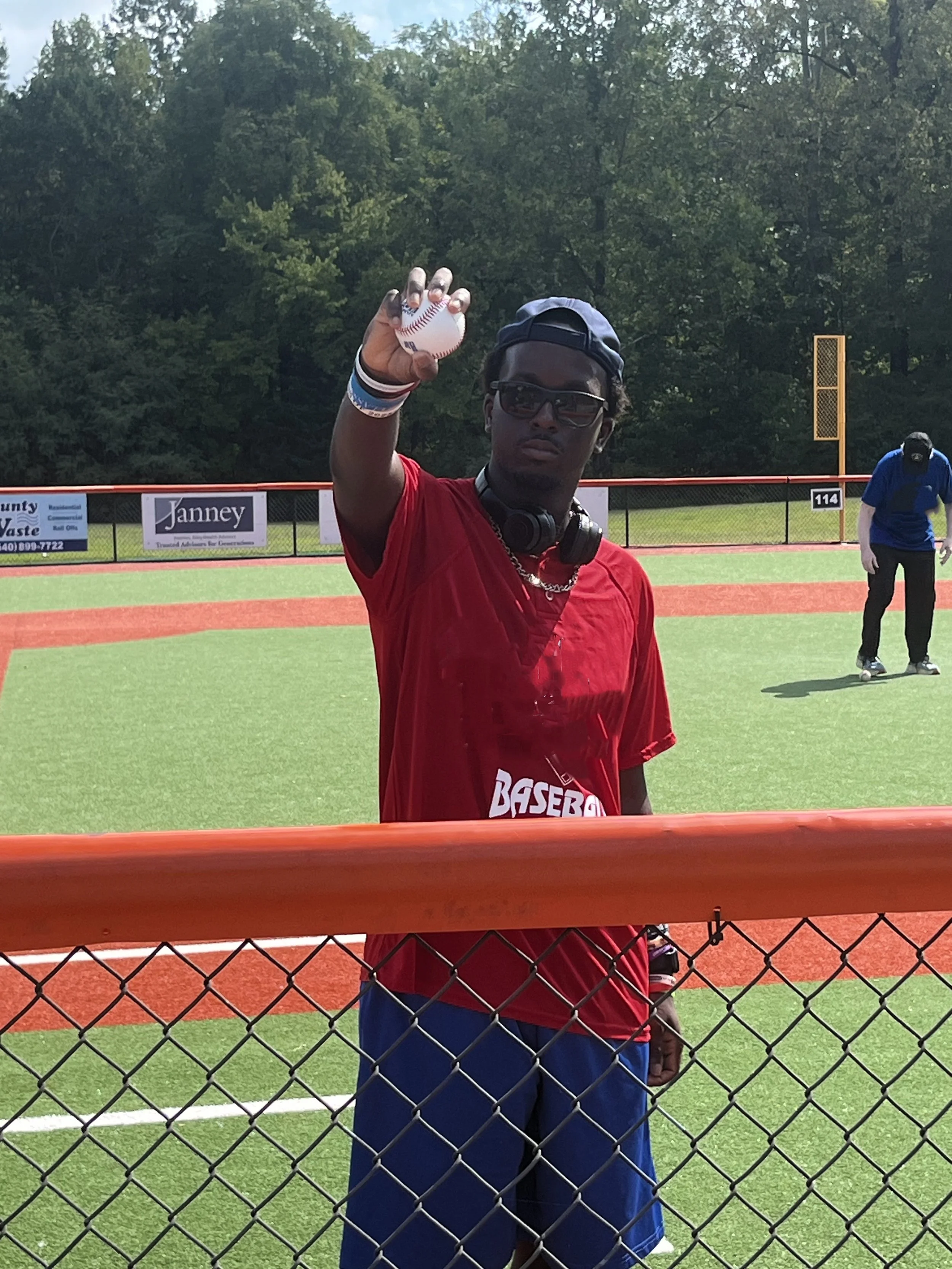A young man in a red shirt and blue shorts holding a baseball in his right hand at a baseball field, with a chain-link fence in the foreground and trees in the background.