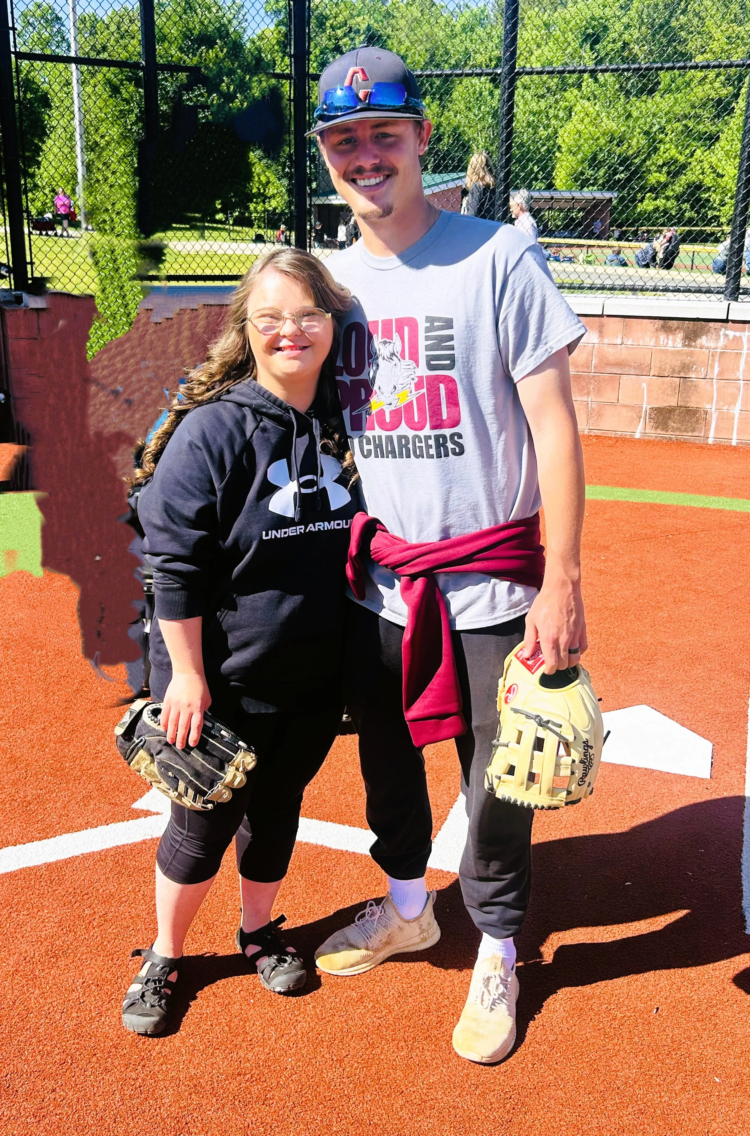 A young man and a young girl posing together on a baseball field. The young man is wearing a gray T-shirt, a red hoodie tied around his waist, gray pants, and holding a baseball glove. The young girl is wearing a black Under Armour hoodie, black capr