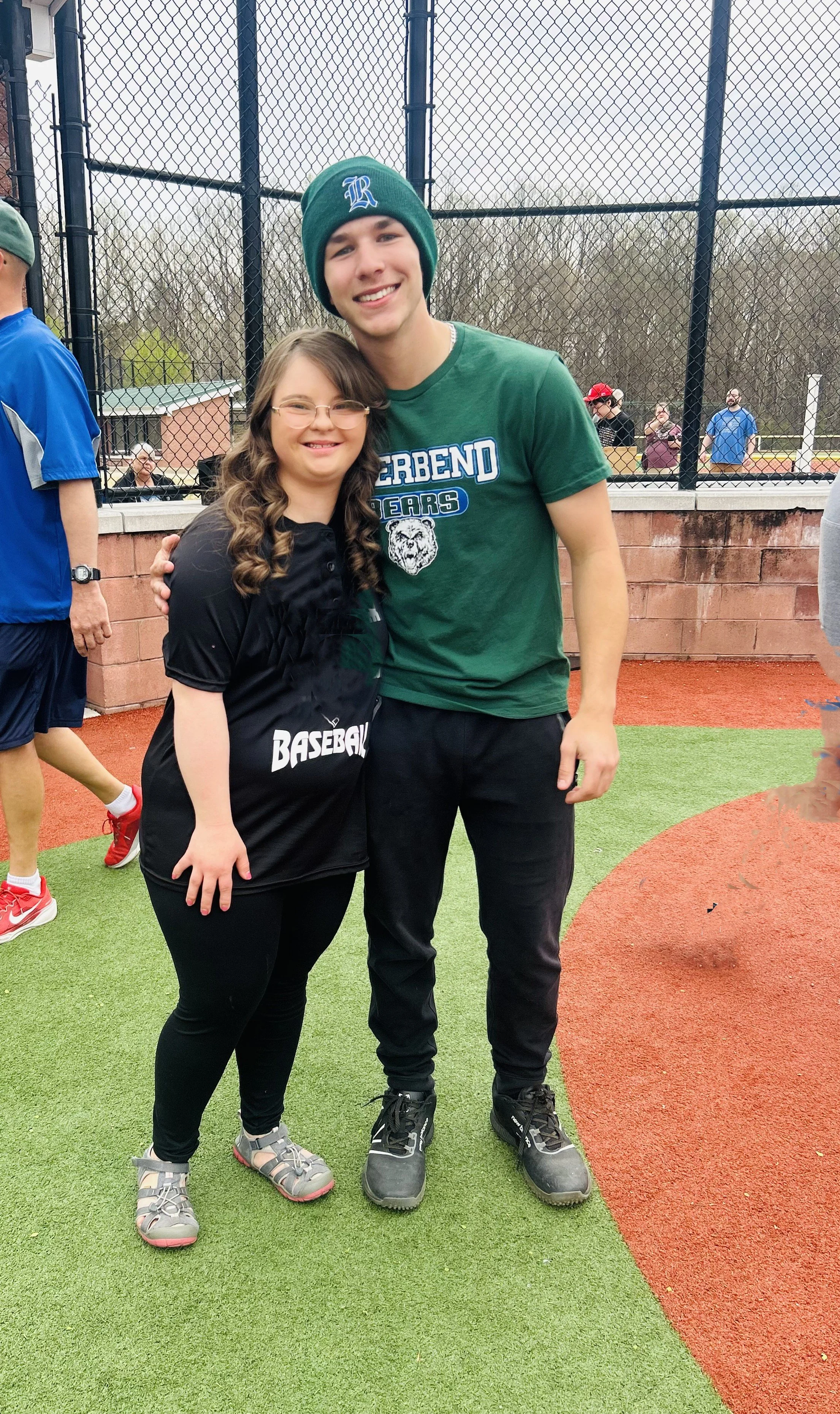 A girl and a young man standing on a baseball field, smiling and posing for a photo. The girl is wearing a black baseball jersey, glasses, and gray sneakers. The young man is wearing a green T-shirt, black pants, black shoes, and a green beanie. A ch