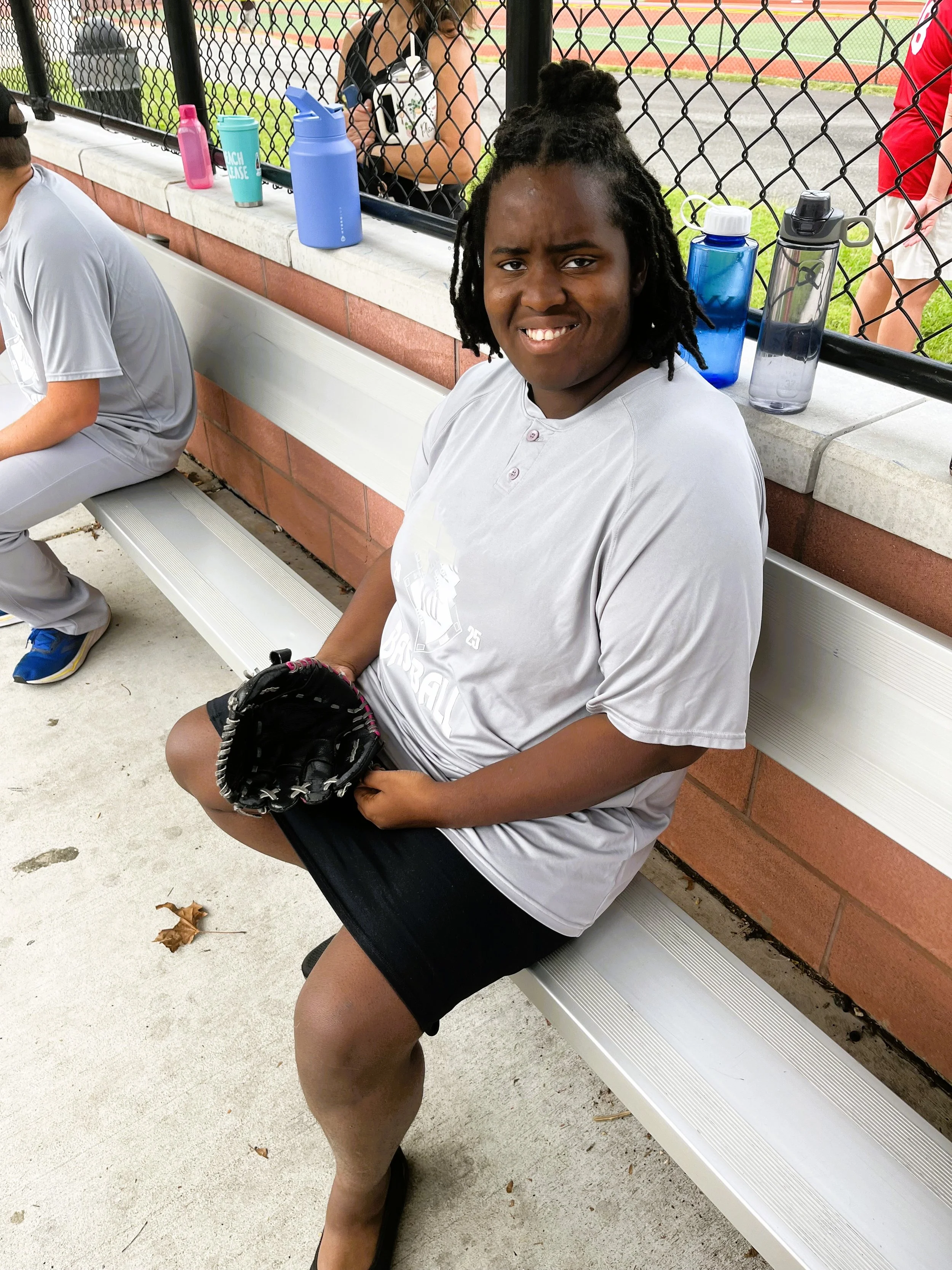 A young woman sitting on a silver bench at a sports field, holding a baseball glove, with water bottles on the ledge behind her, wearing a gray athletic shirt and black shorts.