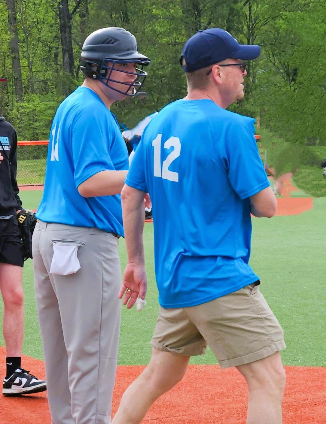 Two men wearing blue shirts numbered 11 and 12 on a baseball field with green trees in the background, one wearing a batting helmet and the other wearing a baseball cap.