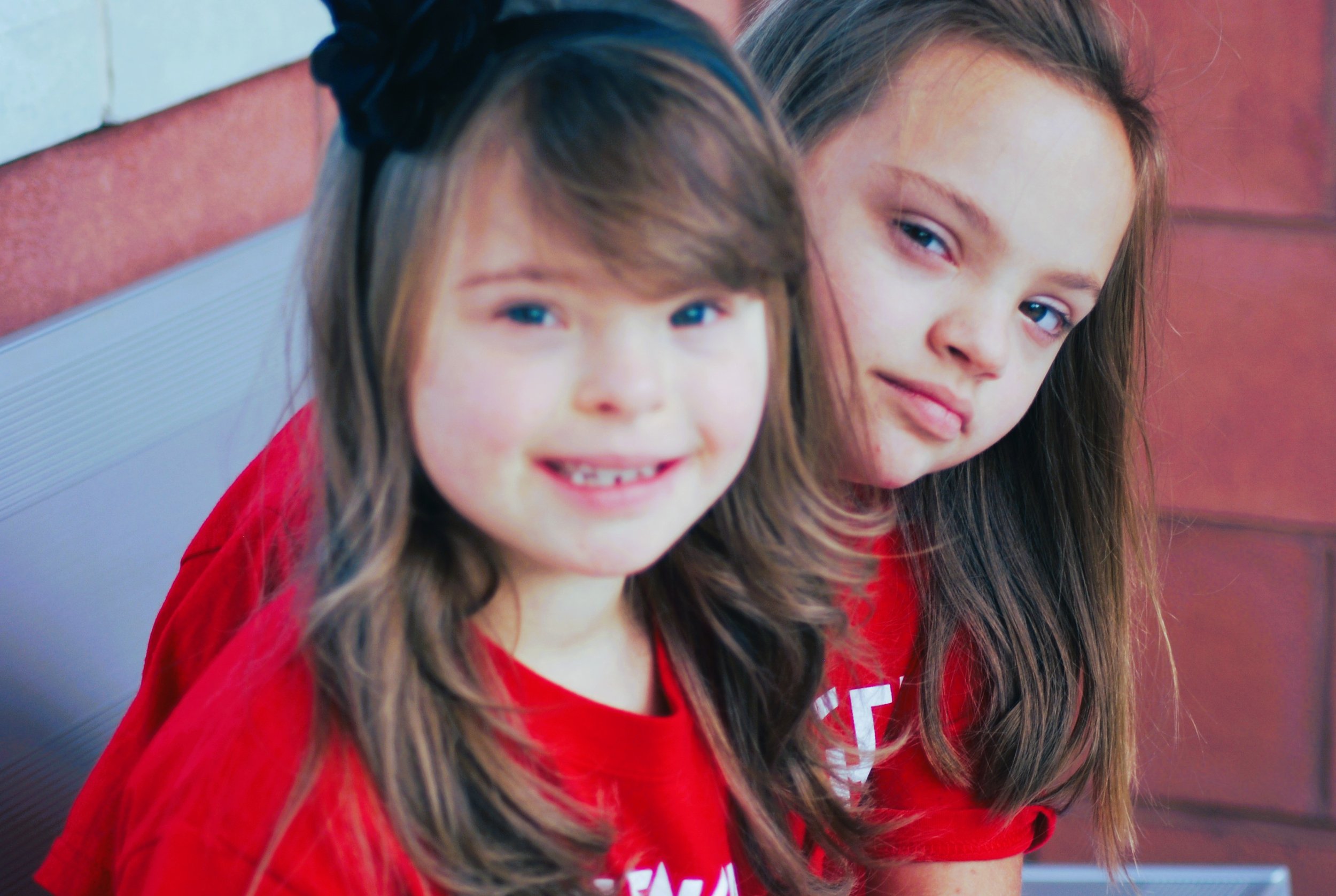 Two young girls sitting next to each other, wearing red shirts, smiling and looking at the camera, outdoors against a brick wall.