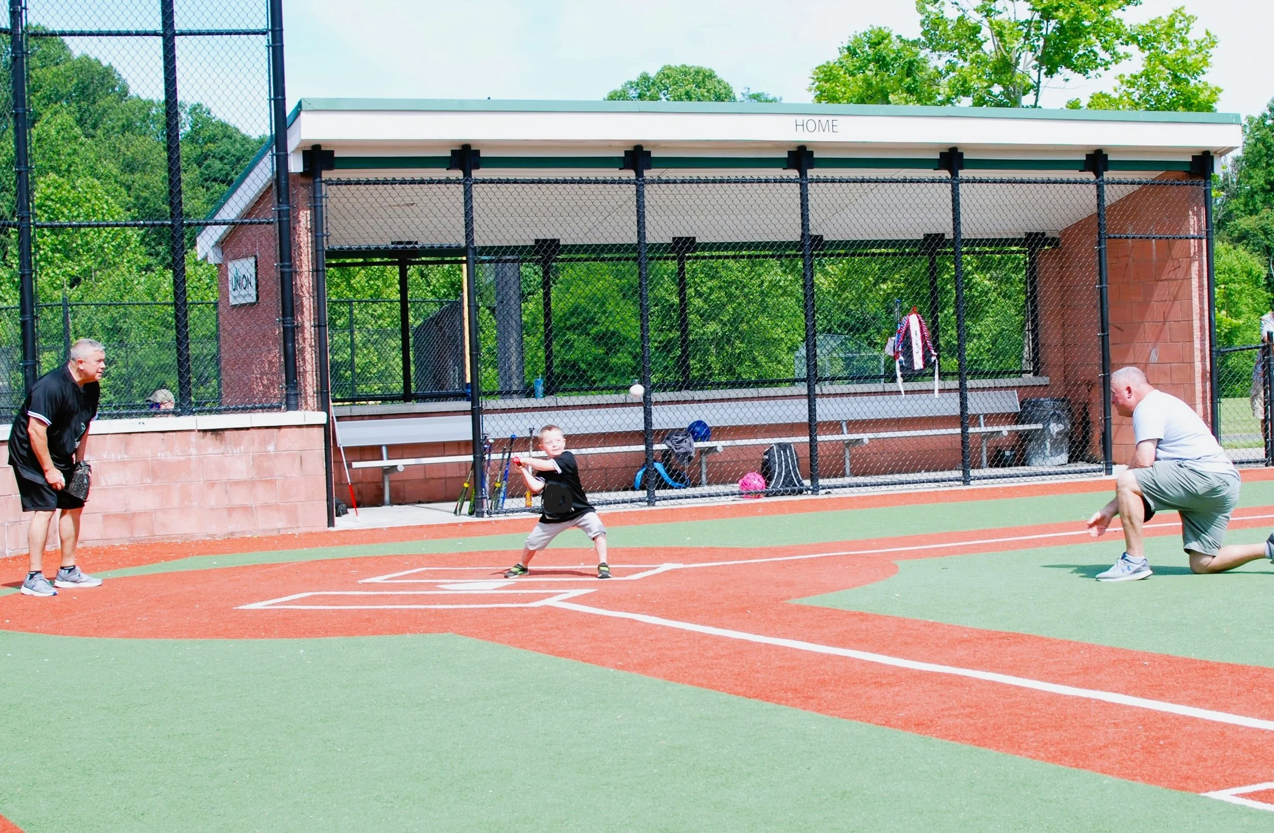 A young boy playing tee ball with two adult men on a baseball field. One man is kneeling and the other man is standing, both ready to catch the ball. The boy is holding a bat, preparing to hit the ball on the tee. The background shows a dugout with s