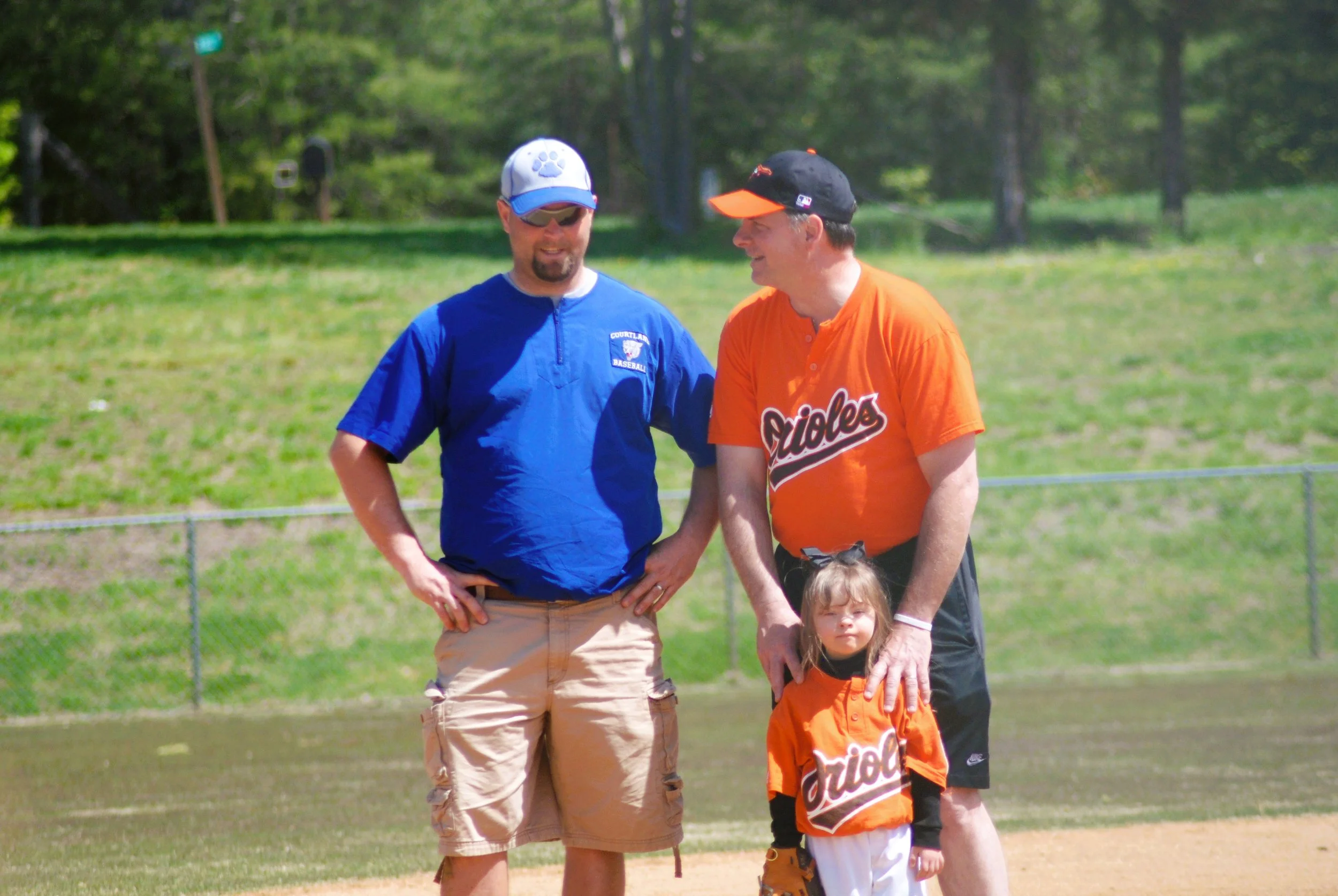Two men and a young girl on a baseball field. One man is wearing a blue shirt and tan shorts, the other man is wearing an orange Baltimore Orioles shirt, and the girl is wearing an orange Orioles jersey. The girl is standing between the two men, who 