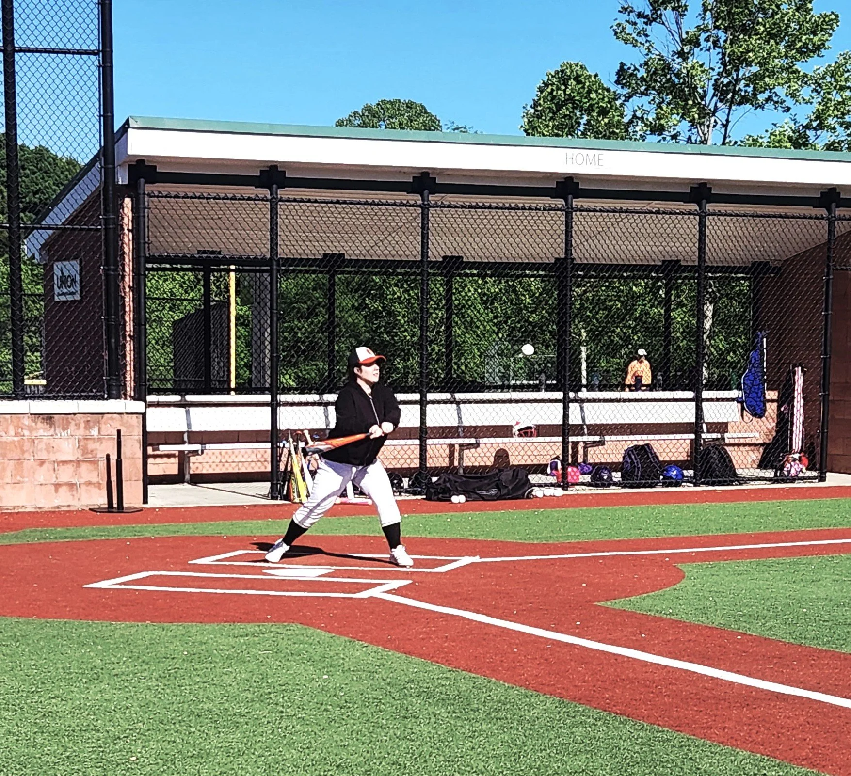 A person standing at home plate preparing to bat in a baseball or softball field, with a chain-link fence, dugout, and trees in the background.
