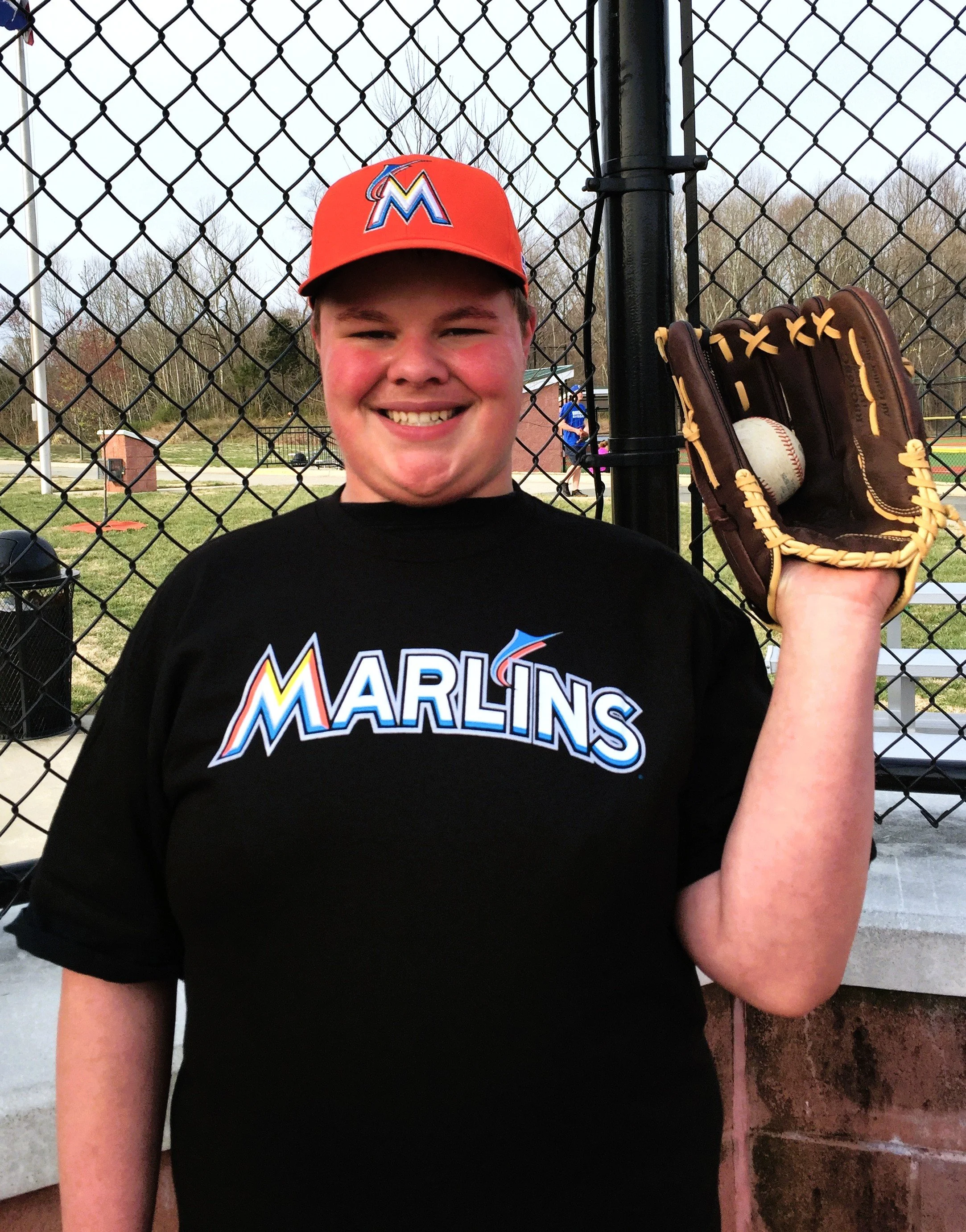 A smiling young male baseball player wearing a Miami Marlins cap and t-shirt holding a baseball glove with a baseball inside at a baseball field.