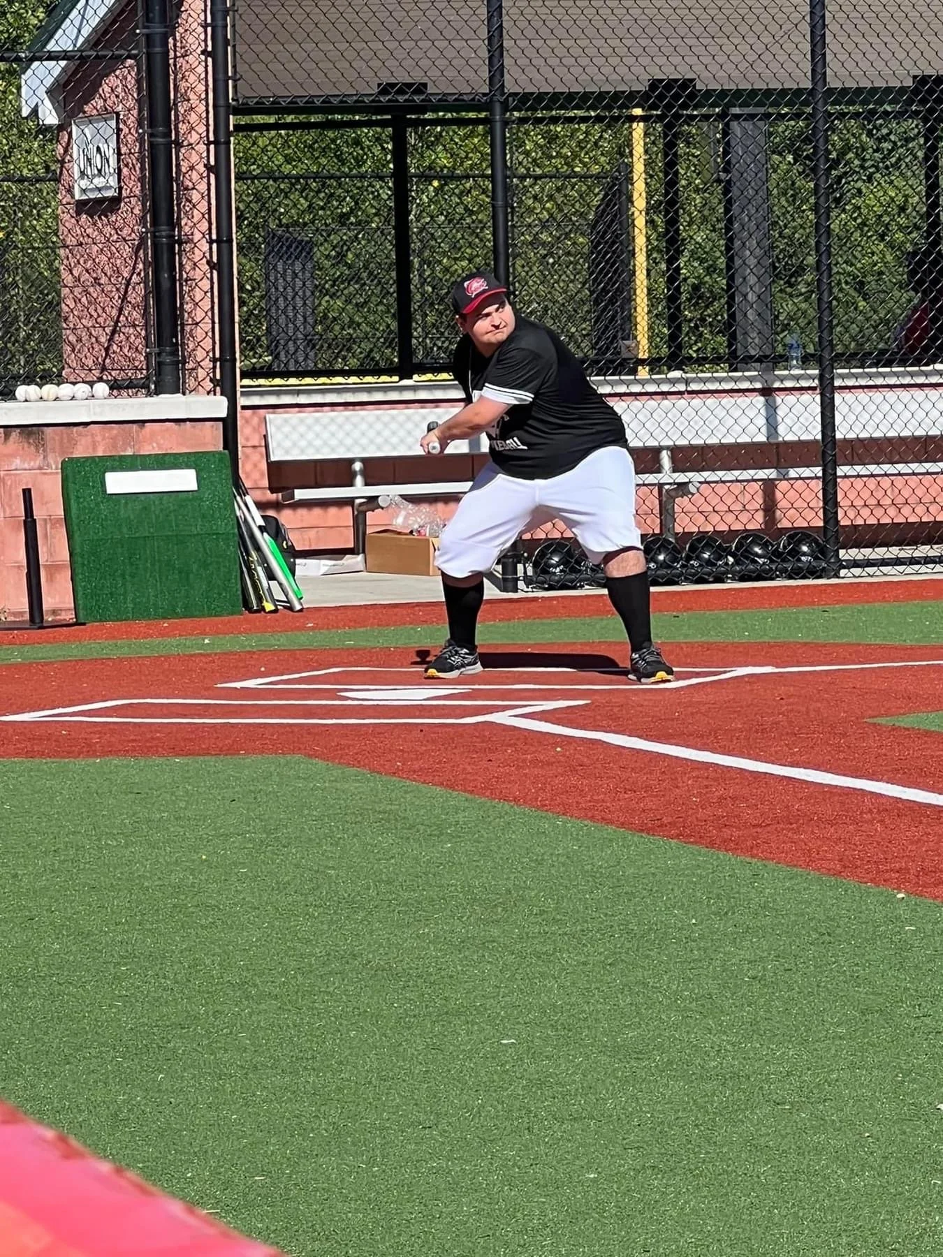 A young man in a black baseball jersey, white pants, and black socks is taking batting practice at a baseball field. He is holding a bat and is standing in a batting stance at home plate. Behind him are baseball equipment and a scoreboard.