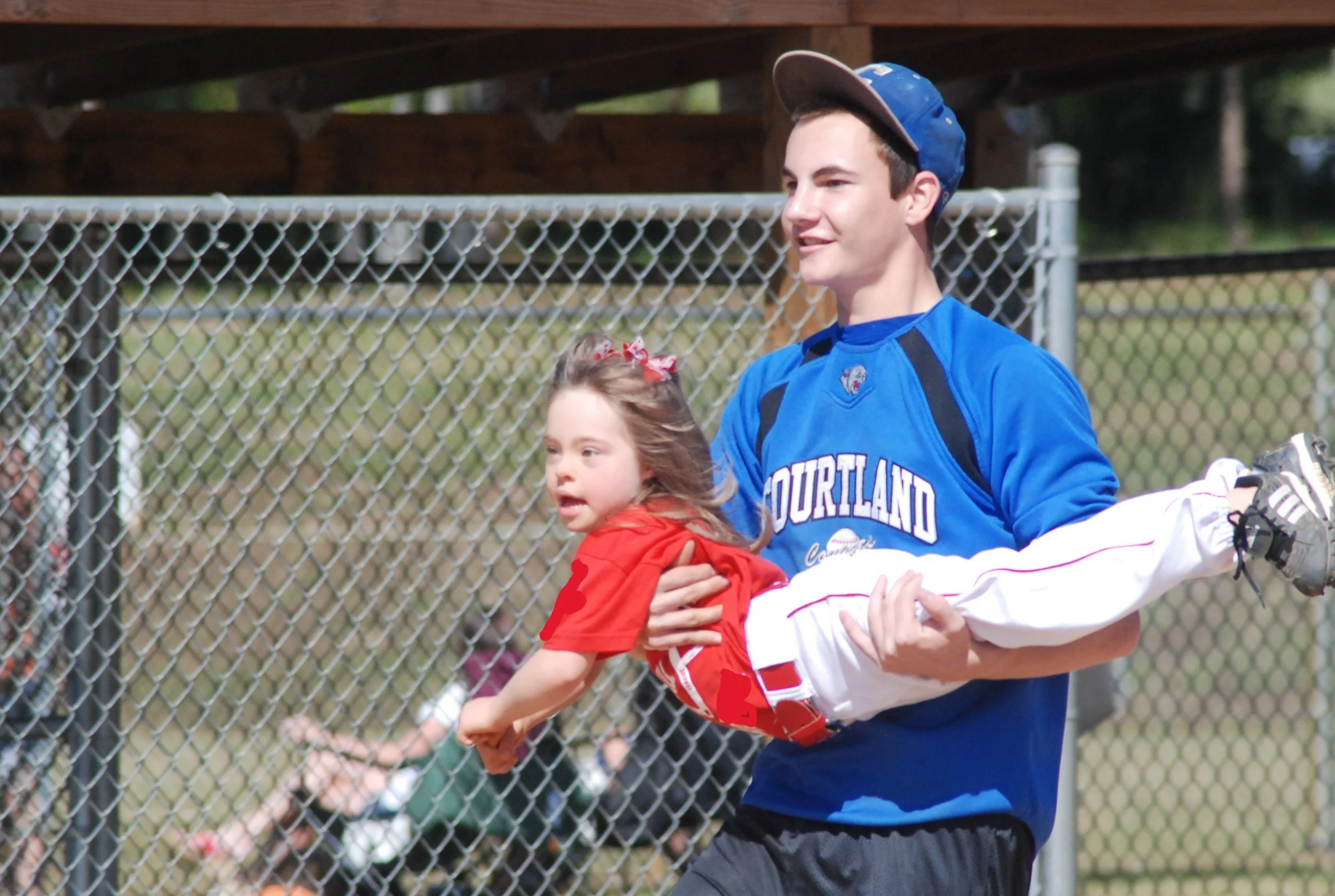 A young man in a blue sports jersey holding a young girl in a red shirt above the ground at a sports field with a chain-link fence in the background.