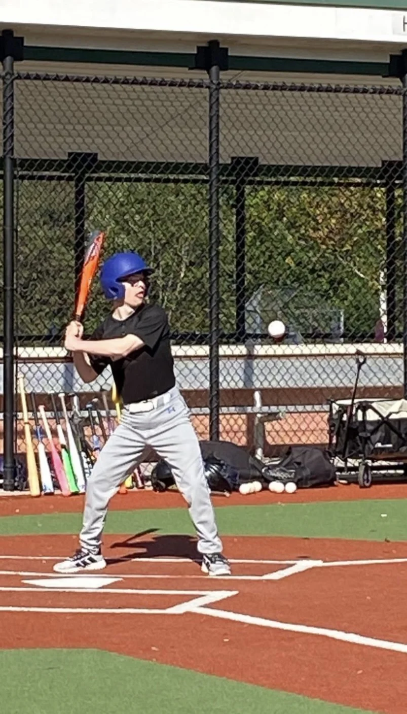 A young baseball player stands at home plate, holding a bat and preparing to swing. The player is wearing a blue helmet, black shirt, gray pants, and sneakers, on a baseball field with a fence and various equipment in the background.
