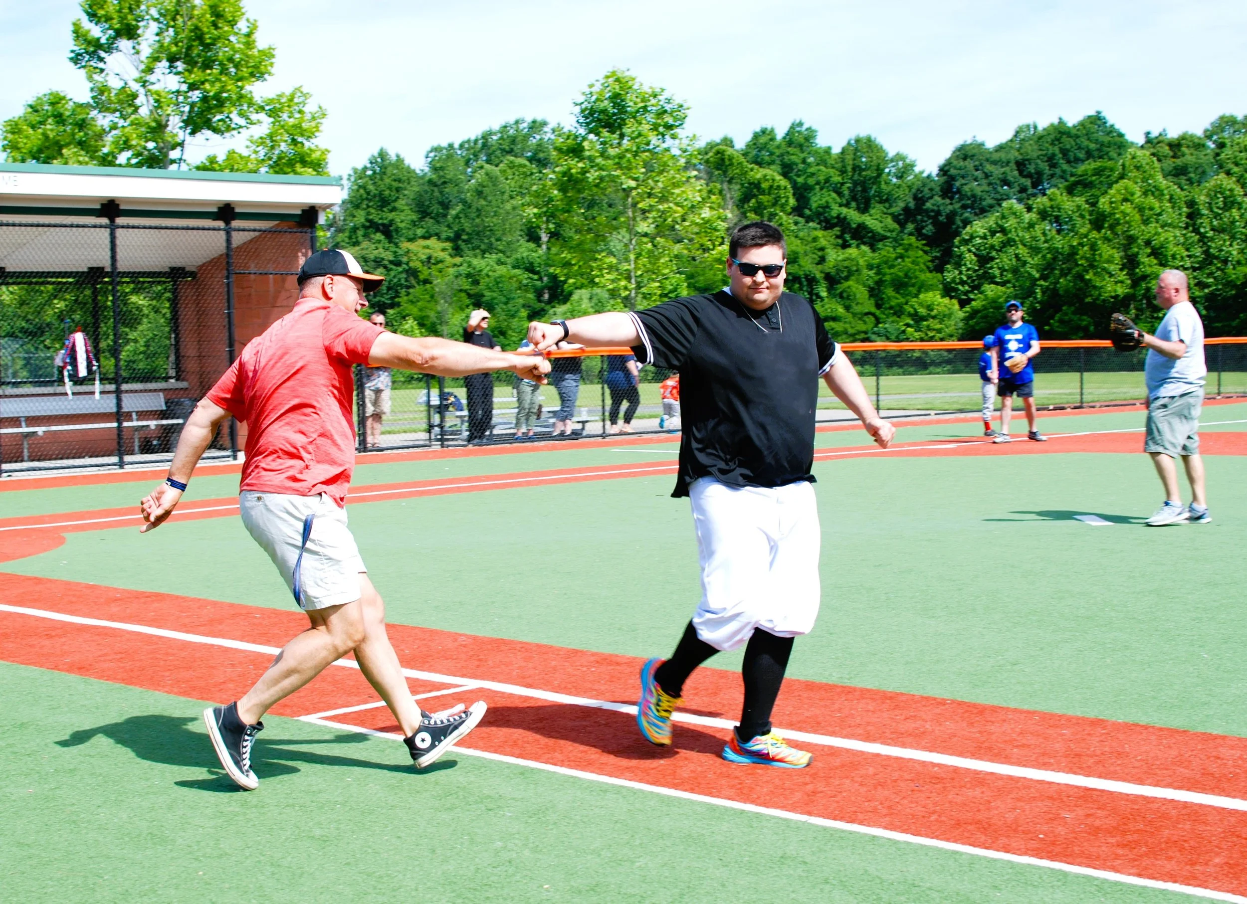 Two men playing catch on a baseball field on a sunny day, one wearing a red shirt and gray shorts, the other wearing a black shirt, white baseball pants, and colorful sneakers, with other people and trees in the background.