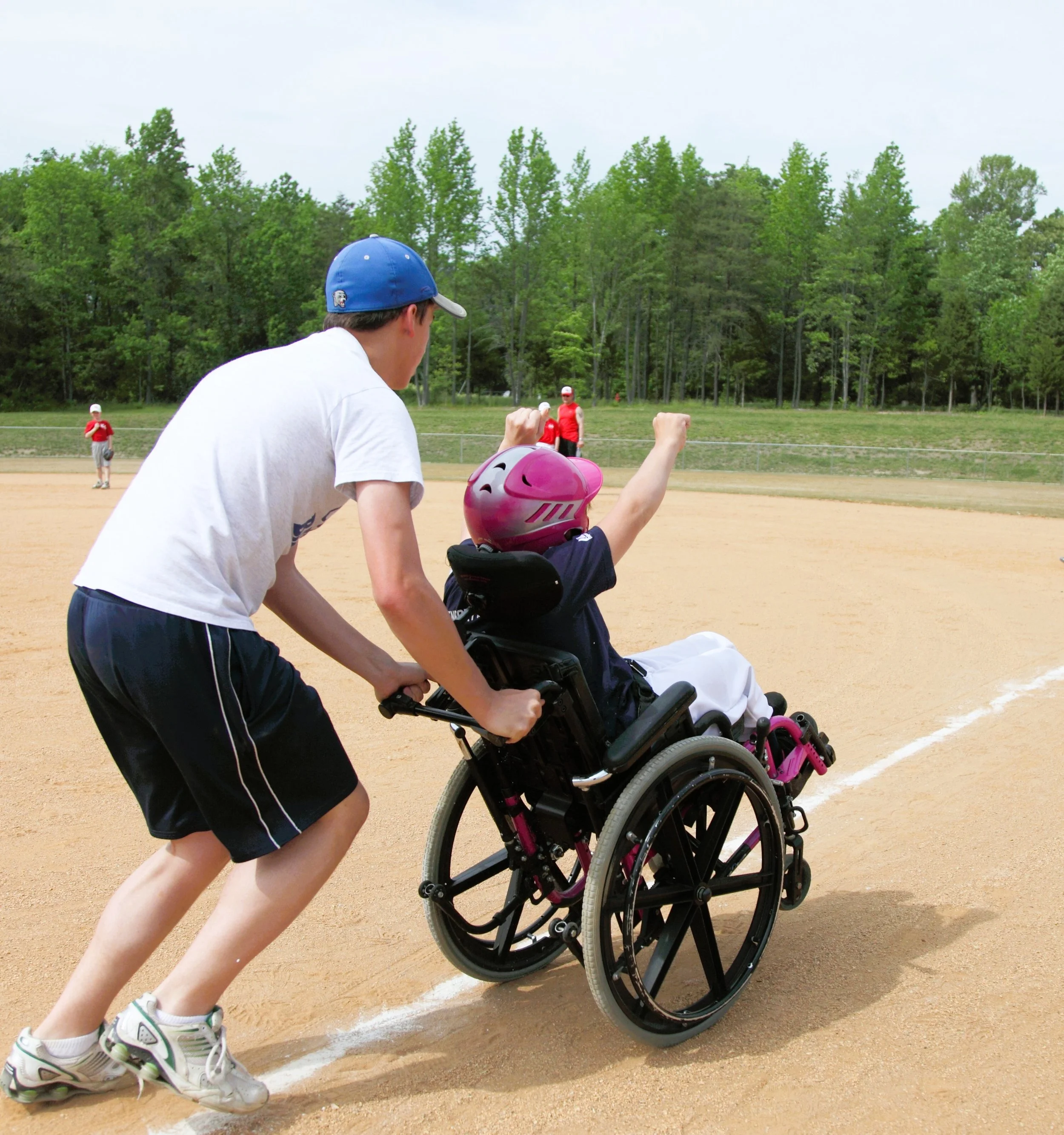 A boy in a wheelchair celebrating as another boy pushes him on a baseball field, with trees and other players in the background.